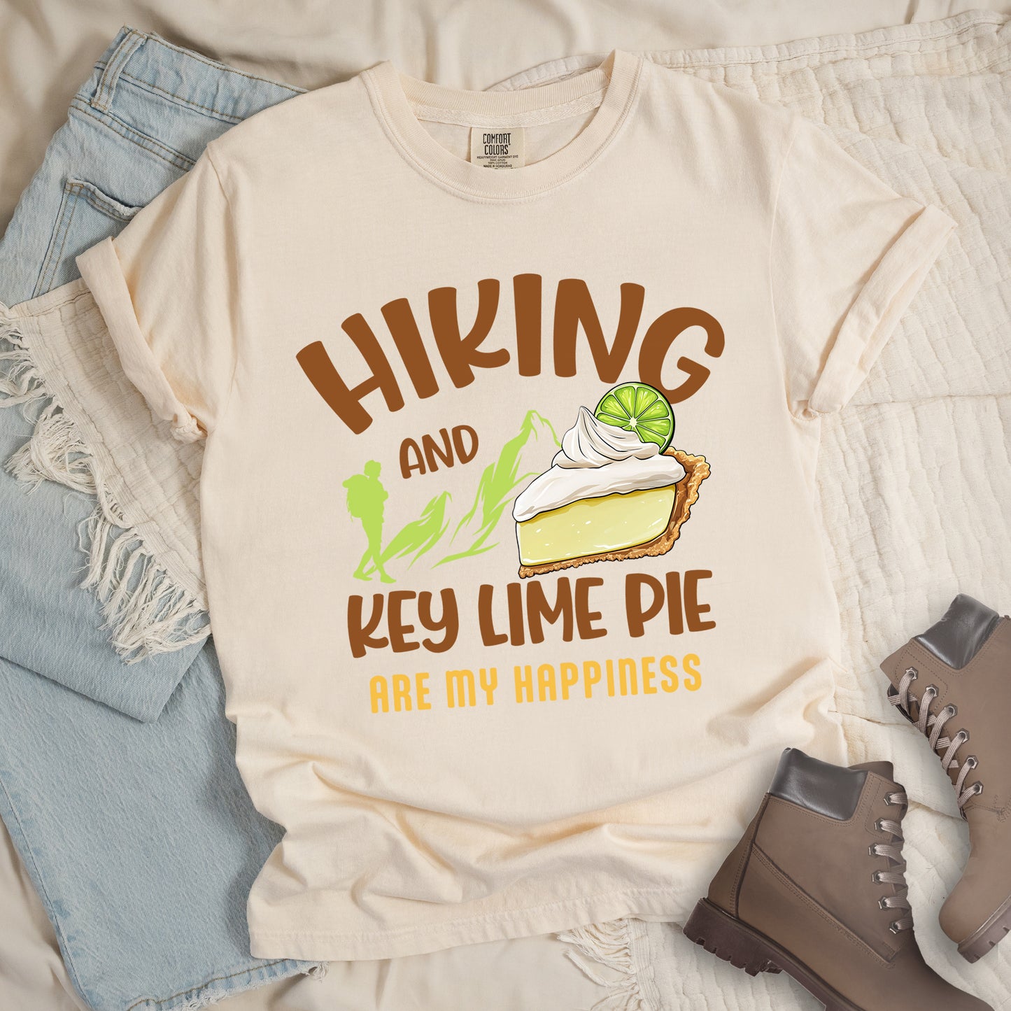 Ivory (Cream White) t-shirt with text "HIKING AND KEY LIME PIE ARE MY HAPPINESS". "HIKING" appears in brown arched letters at the top, "AND" in small brown text, "KEY LIME PIE" in large brown text in the middle, and "ARE MY HAPPINESS" in gold/yellow at the bottom. The design includes green mountain silhouettes with a hiker silhouette and an illustration of a slice of key lime pie with whipped cream and lime garnish in between "AND" and "KEY LIME PIE." 