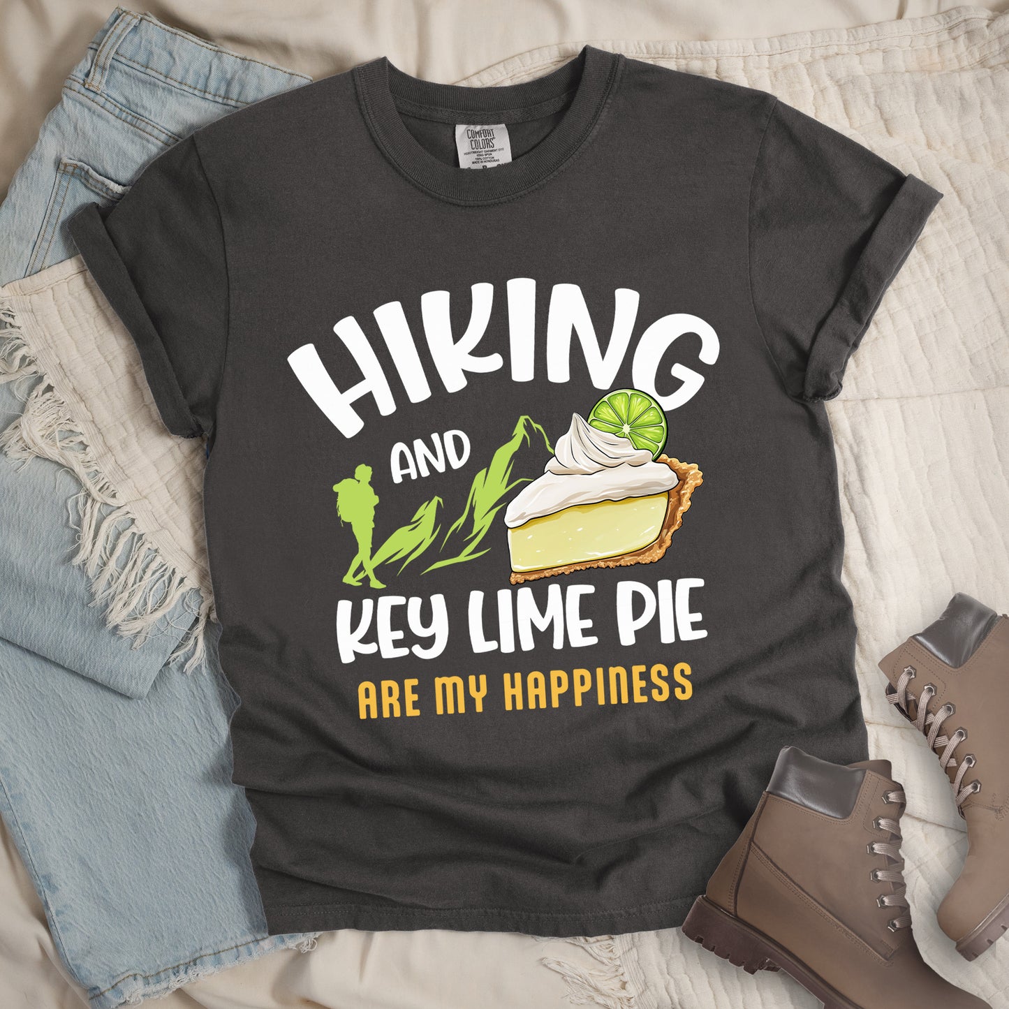 Pepper (Dark Gray) t-shirt with text "HIKING AND KEY LIME PIE ARE MY HAPPINESS". "HIKING" appears in white arched letters at the top, "AND" in small white text, "KEY LIME PIE" in large white text in the middle, and "ARE MY HAPPINESS" in gold/yellow at the bottom. The design includes green mountain silhouettes with a hiker silhouette and an illustration of a slice of key lime pie with whipped cream and lime garnish in between "AND" and "KEY LIME PIE." 