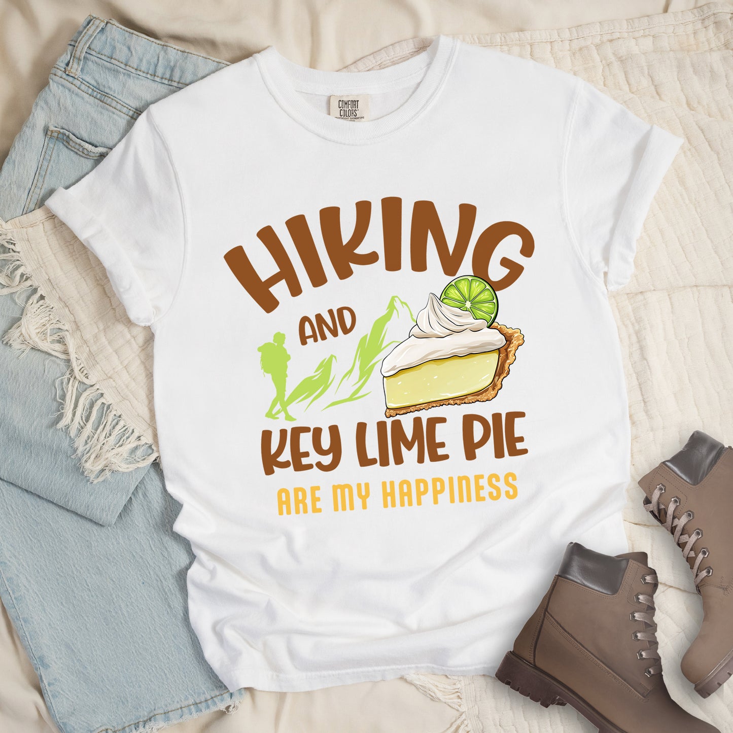 White t-shirt with text "HIKING AND KEY LIME PIE ARE MY HAPPINESS". "HIKING" appears in brown arched letters at the top, "AND" in small brown text, "KEY LIME PIE" in large brown text in the middle, and "ARE MY HAPPINESS" in gold/yellow at the bottom. The design includes green mountain silhouettes with a hiker silhouette and an illustration of a slice of key lime pie with whipped cream and lime garnish in between "AND" and "KEY LIME PIE." 