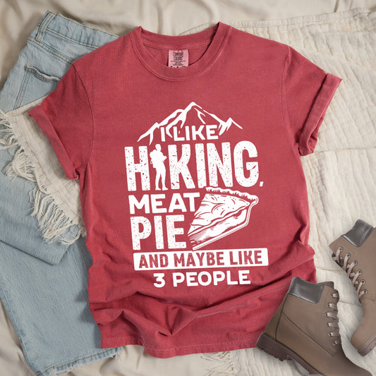 Crimson (Dark Pink) t-shirt with white distressed text reading "I LIKE HIKING, MEAT PIE AND MAYBE LIKE 3 PEOPLE". Mountain silhouettes appear above "I LIKE". The letter "I" in "HIKING" is replaced with a hiker silhouette. Next to "MEAT PIE" is an illustration of a slice of pie. All text and graphics are in white with a weathered, vintage appearance. The design combines outdoor activities, food preferences, and introvert humor. 