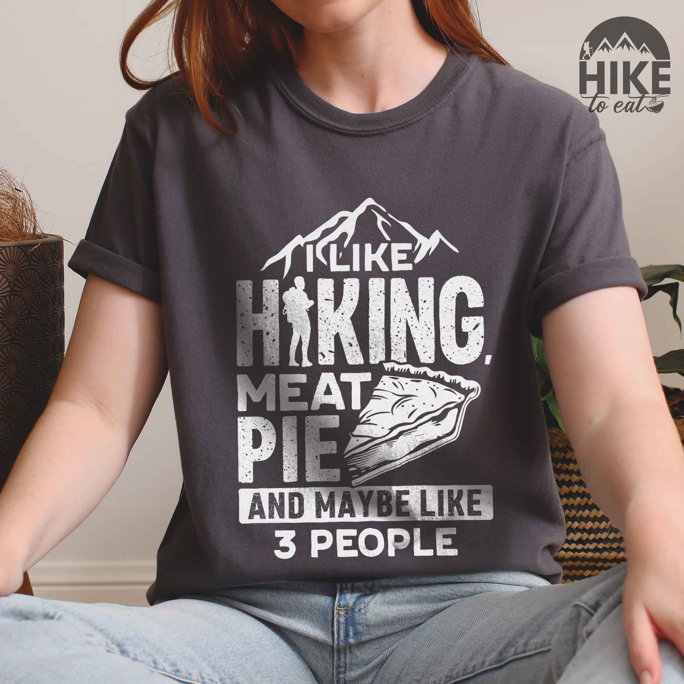 Woman wearing pepper "I like hiking, meat pie and maybe like 3 people" colored vintage tee with bold typography, mountains, birds, illustrated slice of meat pie, and backpacker for pie lovers.