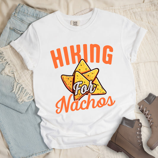 White t-shirt with orange text reading "HIKING For Nachos". "HIKING" appears in large orange letters at the top, "For" in white script across the middle, and "Nachos" in orange script at the bottom. The centerpiece of the design is an illustration of tortilla chips arranged in a star-like pattern, colored yellow-orange with flecks to represent seasonings. The design connects hiking activity with food motivation. 