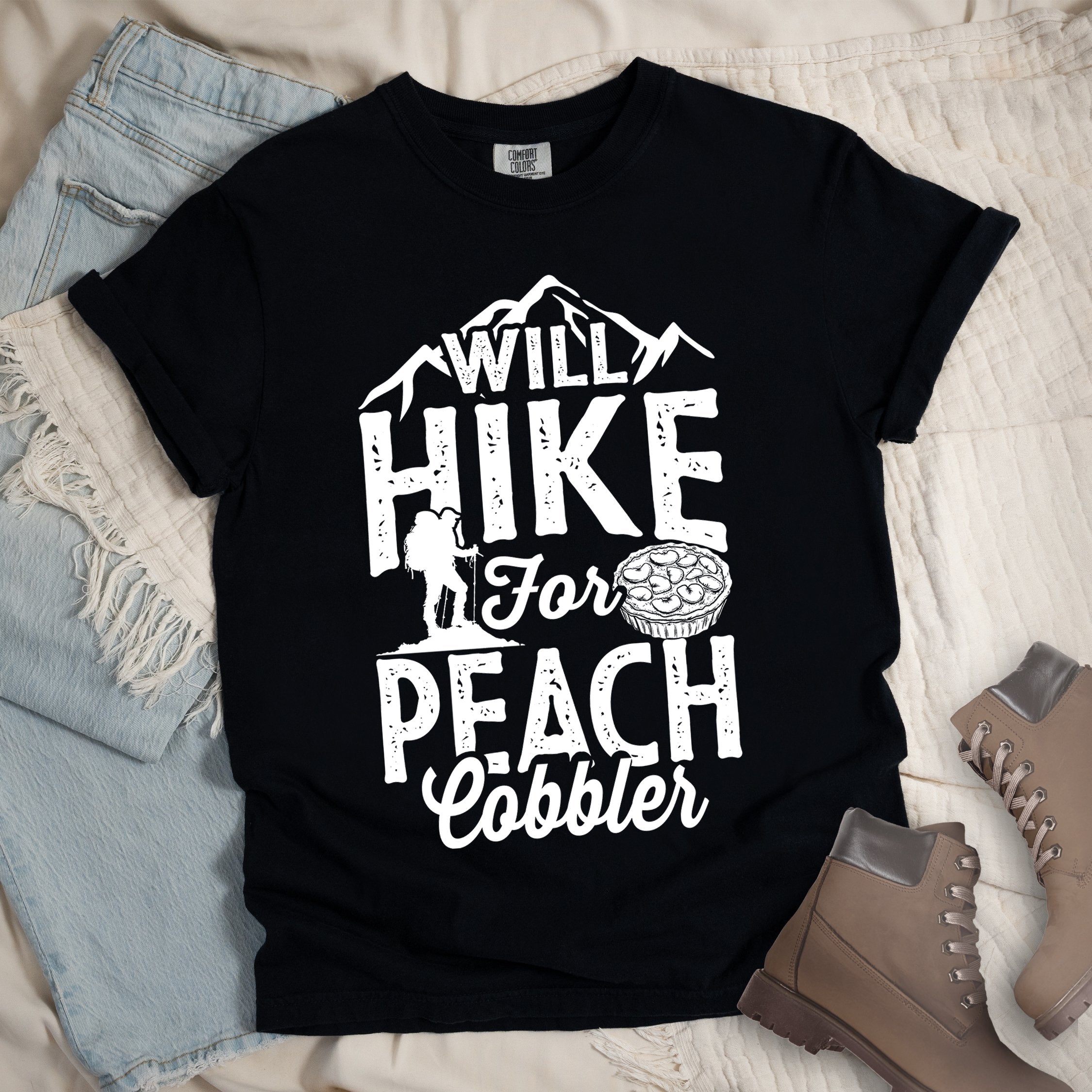 Black shirt with Hiking Design featuring the phrase “Will Hike for Peach Cobbler” and an illustration of a peach cobbler dessert, mountain backdrop, and a hiker silhouette.