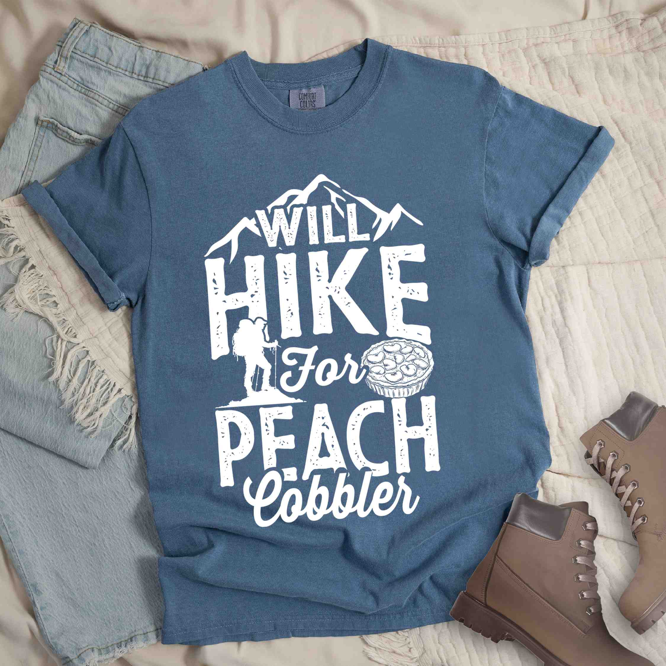 Blue jean "Will hike for peach cobbler" colored vintage tee with bold typography, mountain, birds, illustrated peach cobbler, and backpacker looking for sweet treats.