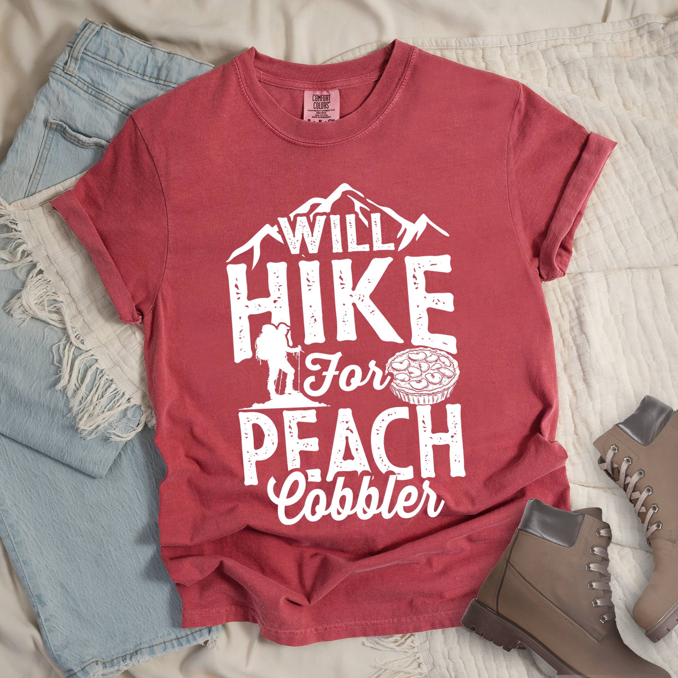 Red shirt with Hiking Design featuring the phrase “Will Hike for Peach Cobbler” and an illustration of a peach cobbler dessert, mountain backdrop, and a hiker silhouette.