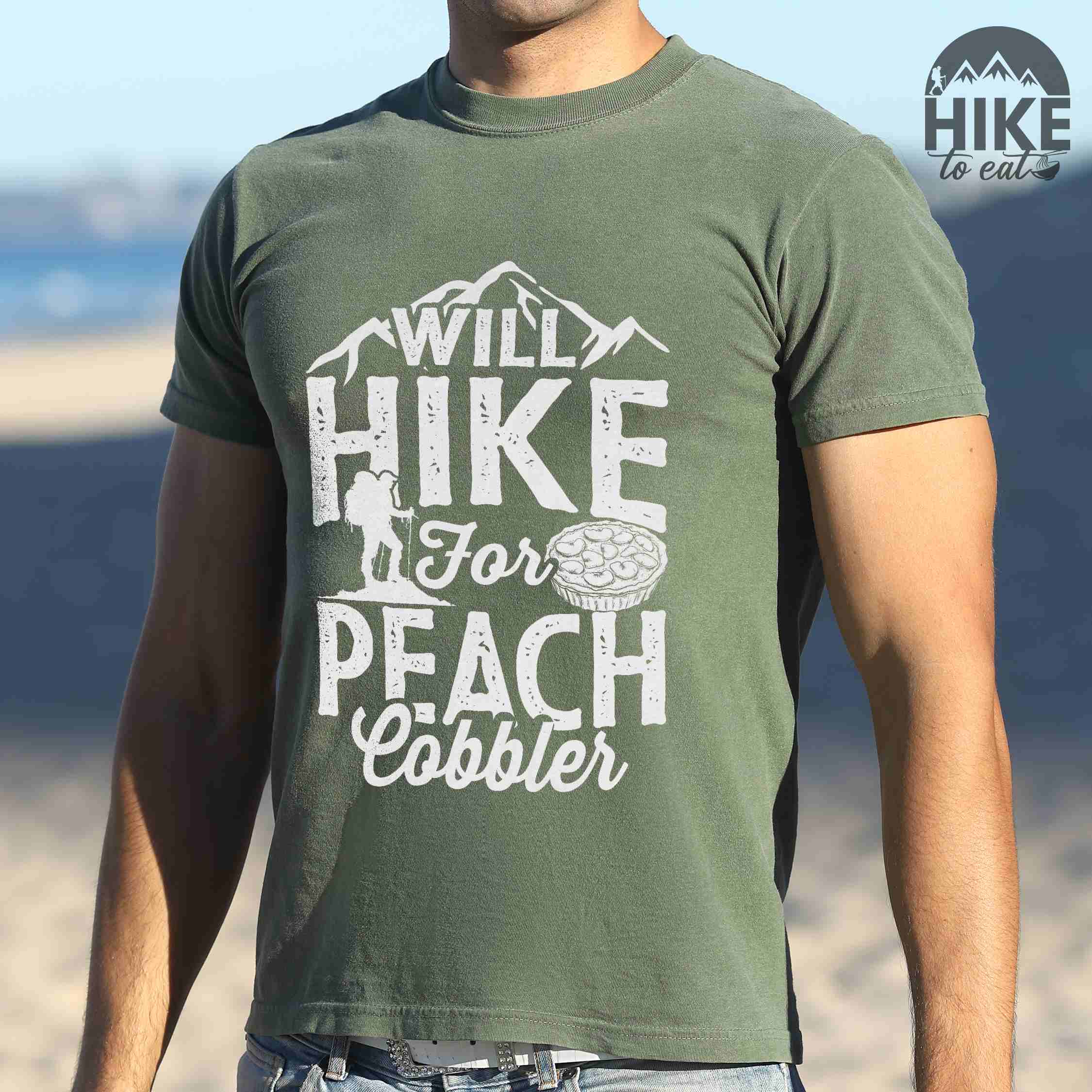 man wearing moss "Will hike for peach cobbler" colored vintage tee with bold typography, mountain, birds, illustrated peach cobbler, and backpacker looking for sweet treats.
