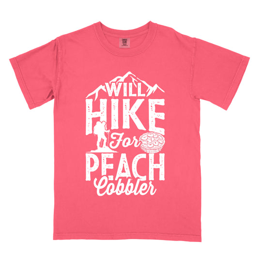 Watermelon (Light Coral) t-shirt with white text reading "WILL HIKE FOR PEACH COBBLER". "WILL HIKE" appears in large distressed letters at the top with mountain silhouettes, "For" in white script across the middle, and "PEACH COBBLER" in large distressed letters at the bottom. The design includes a small hiker figure with backpack on the left and an illustration of a peach cobbler pie. The design humorously connects hiking activity with dessert motivation.