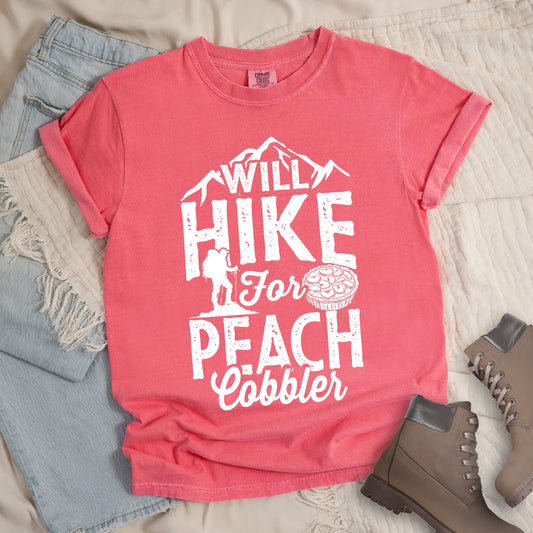 Watermelon (Light Coral) t-shirt with white text reading "WILL HIKE FOR PEACH COBBLER". "WILL HIKE" appears in large distressed letters at the top with mountain silhouettes, "For" in white script across the middle, and "PEACH COBBLER" in large distressed letters at the bottom. The design includes a small hiker figure with backpack on the left and an illustration of a peach cobbler pie. The design humorously connects hiking activity with dessert motivation. 