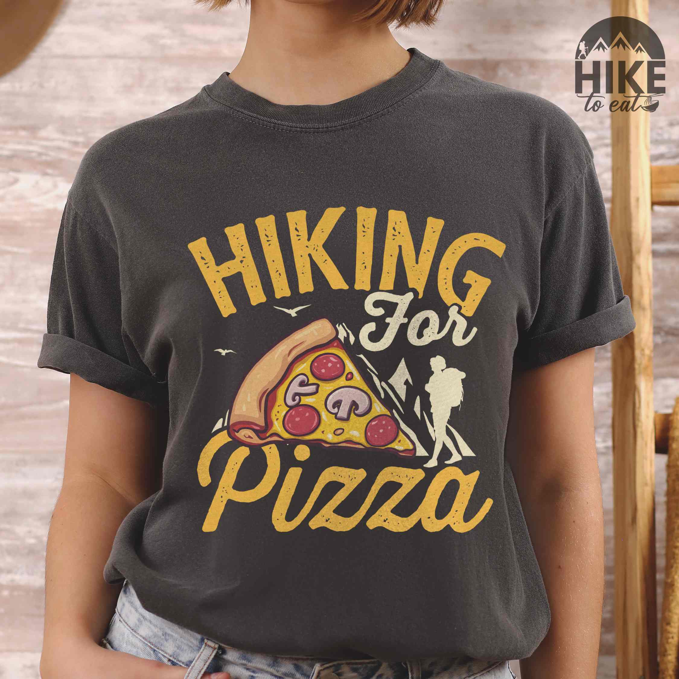 Woman wearing pepper "Hiking for Pizza" colored vintage tee with bold typography, birds, mountains, illustrated pizza slice, and backpacker for pizza lovers.