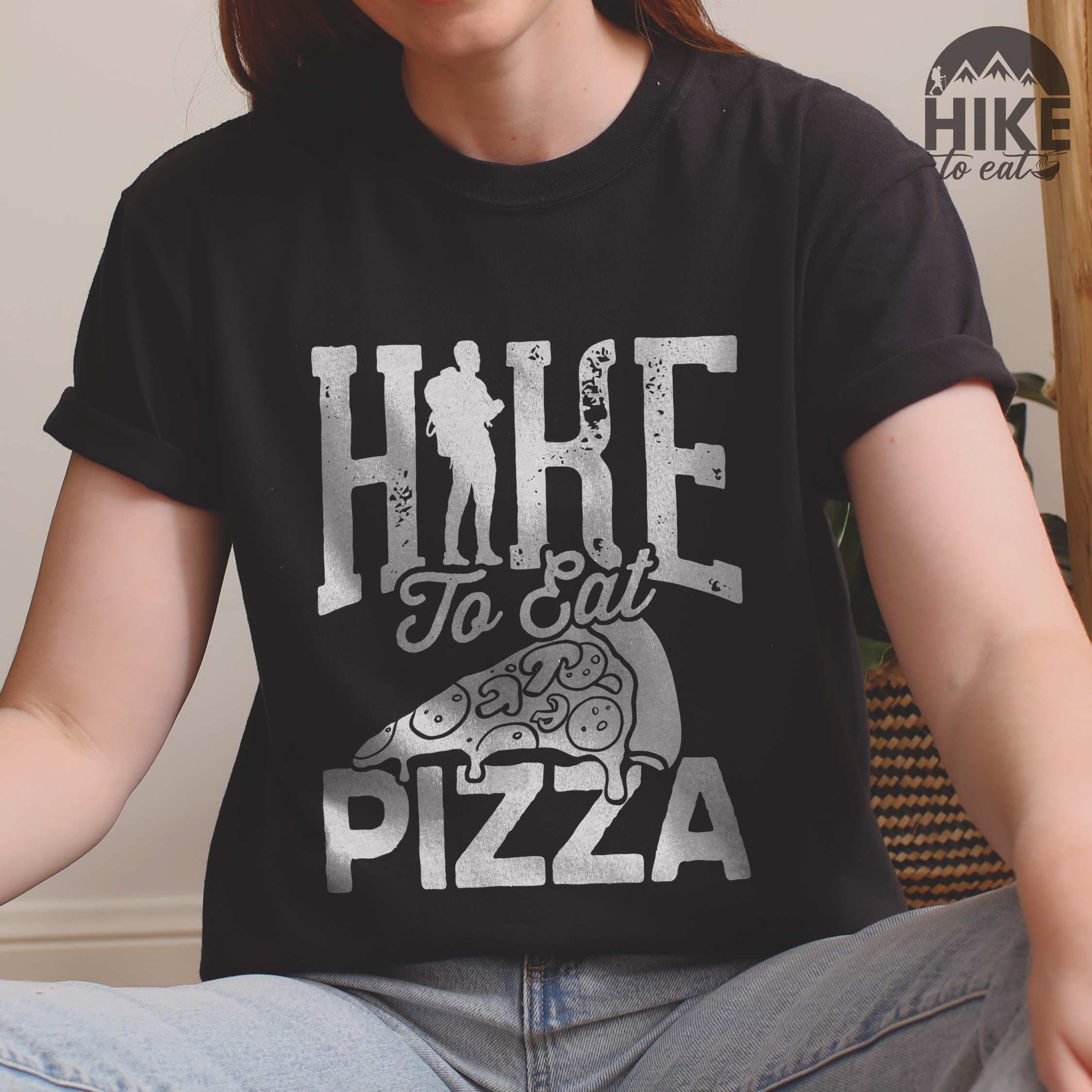 woman wearing black "Hike to eat Pizza" colored vintage tee with bold typography, birds, mountains, illustrated pizza slice, and backpacker for pizza lover.