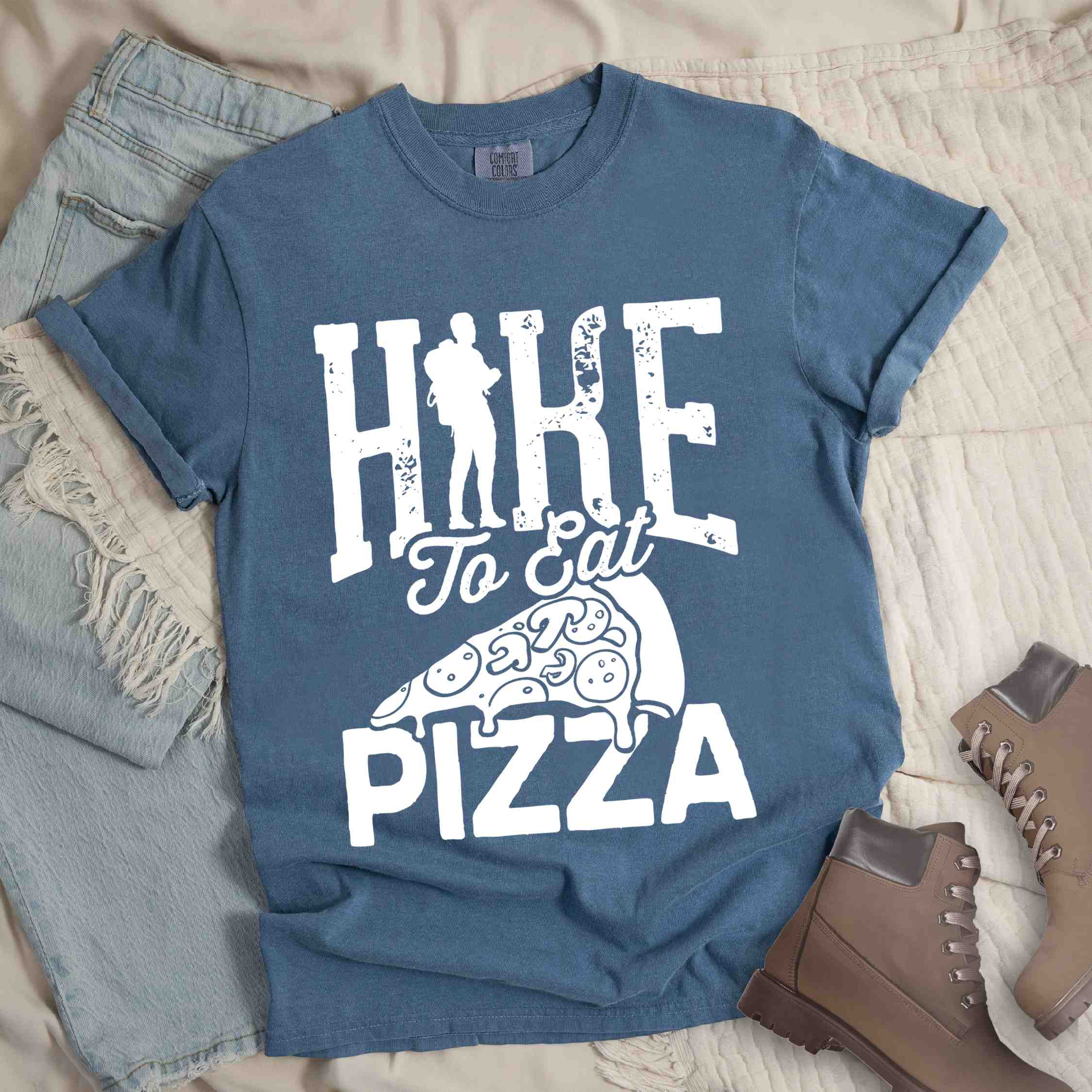 Blue jean "Hike to eat Pizza" colored vintage tee with bold typography, birds, mountains, illustrated pizza slice, and backpacker for pizza lover.