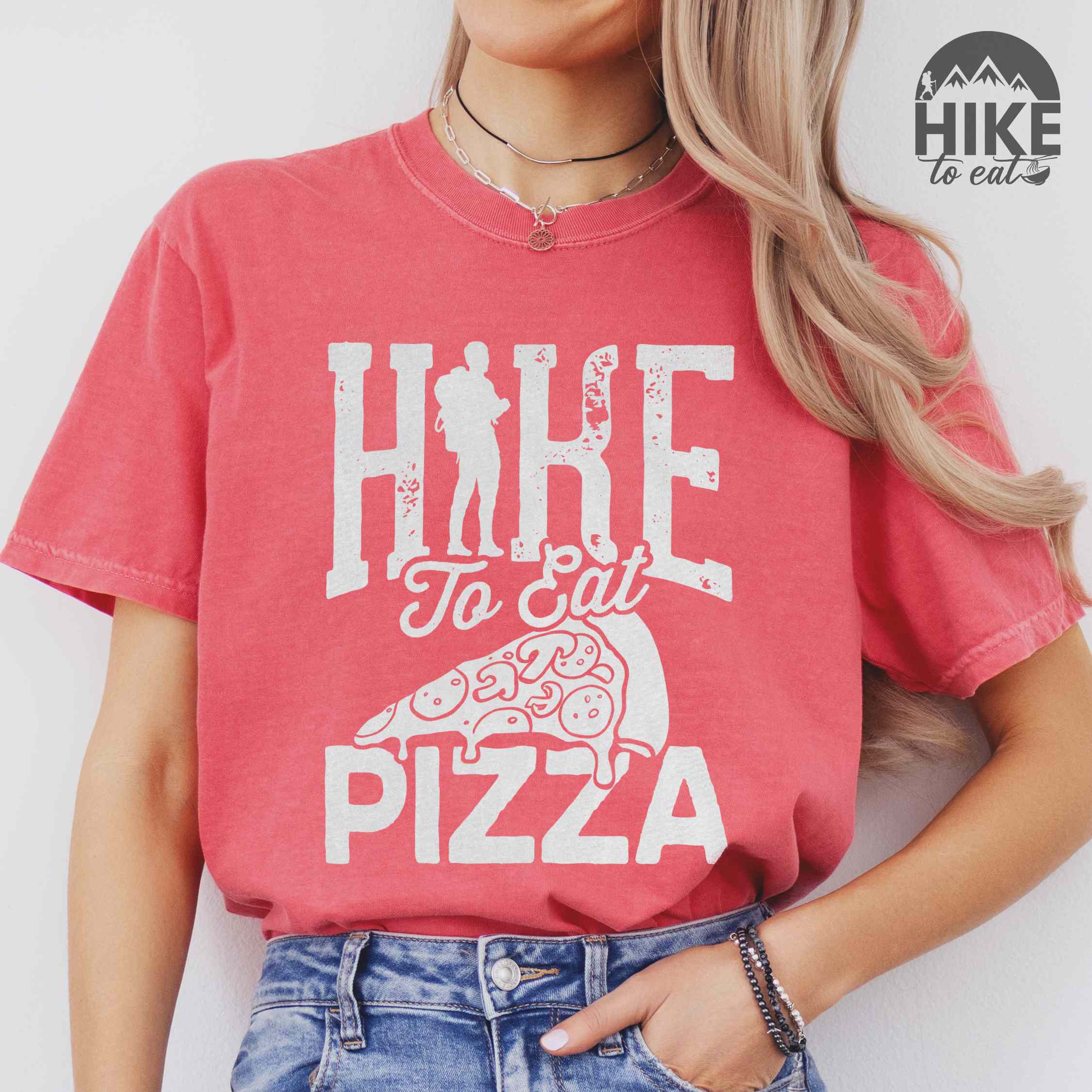 woman wearing watermelon "Hike to eat Pizza" colored vintage tee with bold typography, birds, mountains, illustrated pizza slice, and backpacker for pizza lover.
