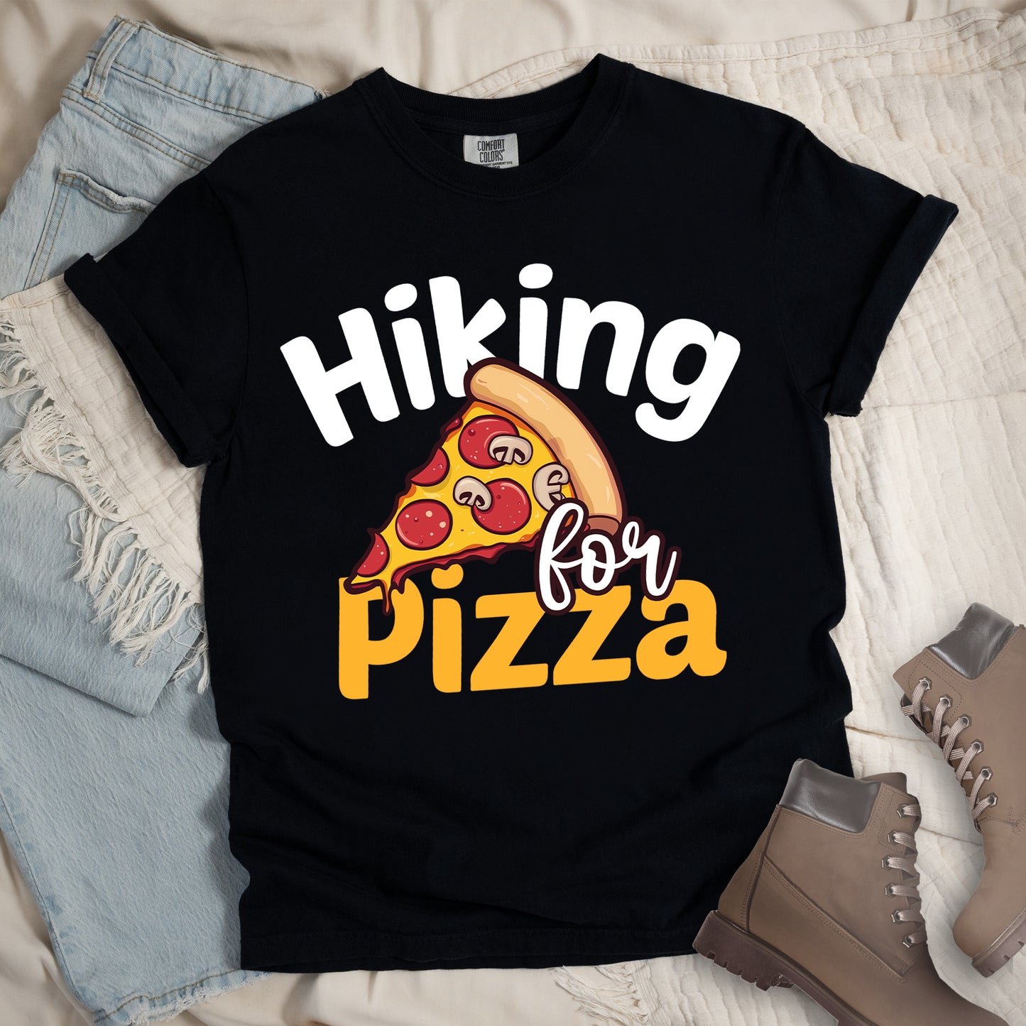 Black t-shirt with white and orange text reading "Hiking for PIZZA". "Hiking" appears in white curved text at the top, "for" in small white script in the middle, and "PIZZA" in bold orange letters at the bottom. The centerpiece is a detailed illustration of a pizza slice with pepperoni and mushroom toppings on a yellow cheese background with tan crust. Features a classic crew neck and casual rolled sleeves, styled alongside light wash denim and brown hiking boots. 