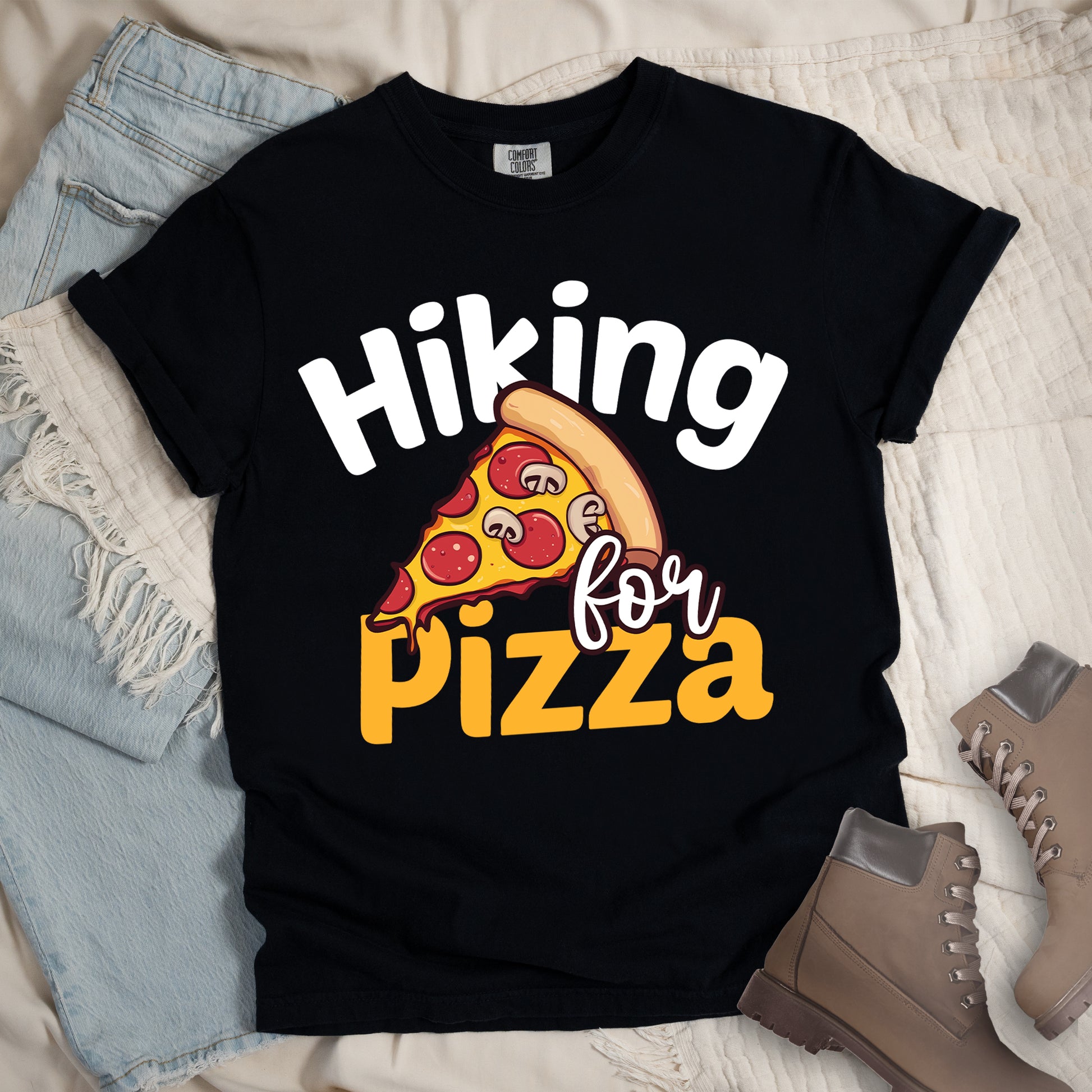 Black t-shirt with white and orange text reading "Hiking for PIZZA". "Hiking" appears in white curved text at the top, "for" in small white script in the middle, and "PIZZA" in bold orange letters at the bottom. The centerpiece is a detailed illustration of a pizza slice with pepperoni and mushroom toppings on a yellow cheese background with tan crust. Features a classic crew neck and casual rolled sleeves, styled alongside light wash denim and brown hiking boots. 