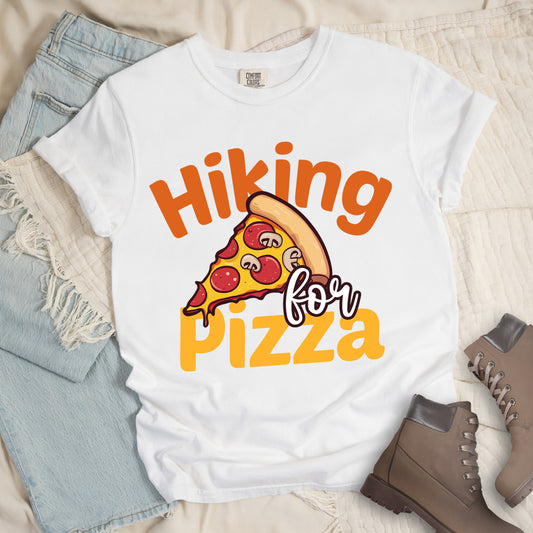 White t-shirt with yellow and orange text reading "Hiking for PIZZA". "Hiking" appears in orange curved text at the top, "for" in small white script in the middle, and "PIZZA" in bold yellow letters at the bottom. The centerpiece is a detailed illustration of a pizza slice with pepperoni and mushroom toppings on a yellow cheese background with tan crust. Features a classic crew neck and casual rolled sleeves, styled alongside light wash denim and brown hiking boots. 