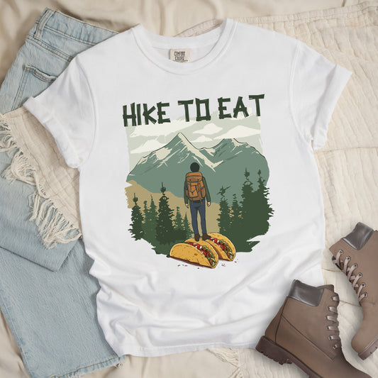 White t-shirt with "HIKE TO EAT" printed at the top in dark green text. The main graphic shows a hiker with backpack standing on two tacos, facing a scenic mountain landscape with pine trees in the foreground and snow-capped peaks in the background. The shirt features a classic crew neck and casual rolled sleeves, styled alongside light wash denim and brown hiking boots.