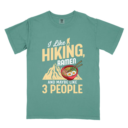 Light Green t-shirt with mixed text reading "I Like HIKING, RAMEN AND MAYBE LIKE 3 PEOPLE". "I Like" in white script at top with small gold hiker silhouette, "HIKING," in large gold letters, "RAMEN" in white, and "AND MAYBE LIKE 3 PEOPLE" in white and gold. Features a colorful illustration of a red ramen bowl with eggs, noodles and chopsticks, positioned above a gold mountain silhouette.