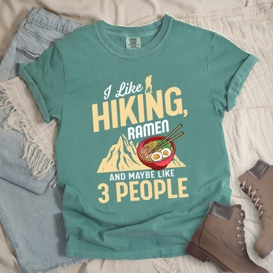 Light Green t-shirt with mixed text reading "I Like HIKING, RAMEN AND MAYBE LIKE 3 PEOPLE". "I Like" in white script at top with small gold hiker silhouette, "HIKING," in large gold letters, "RAMEN" in white, and "AND MAYBE LIKE 3 PEOPLE" in white and gold. Features a colorful illustration of a red ramen bowl with eggs, noodles and chopsticks, positioned above a gold mountain silhouette. Features a classic crew neck and casual rolled sleeves, styled alongside light wash denim and brown hiking boots. 