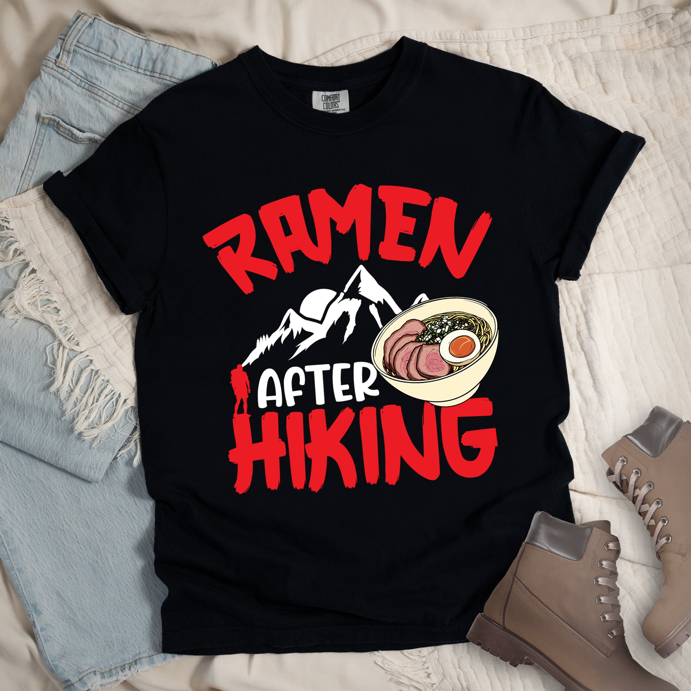 Black t-shirt with red text reading "RAMEN AFTER HIKING". "RAMEN" appears in bold red letters at the top, "AFTER" in white in the middle, and "HIKING" in bold red at the bottom. Features white mountain silhouettes in the center with a red hiker figure, and a detailed illustration of a ramen bowl with sliced meat, egg, and green onions on the right. The design connects outdoor activities with food rewards.