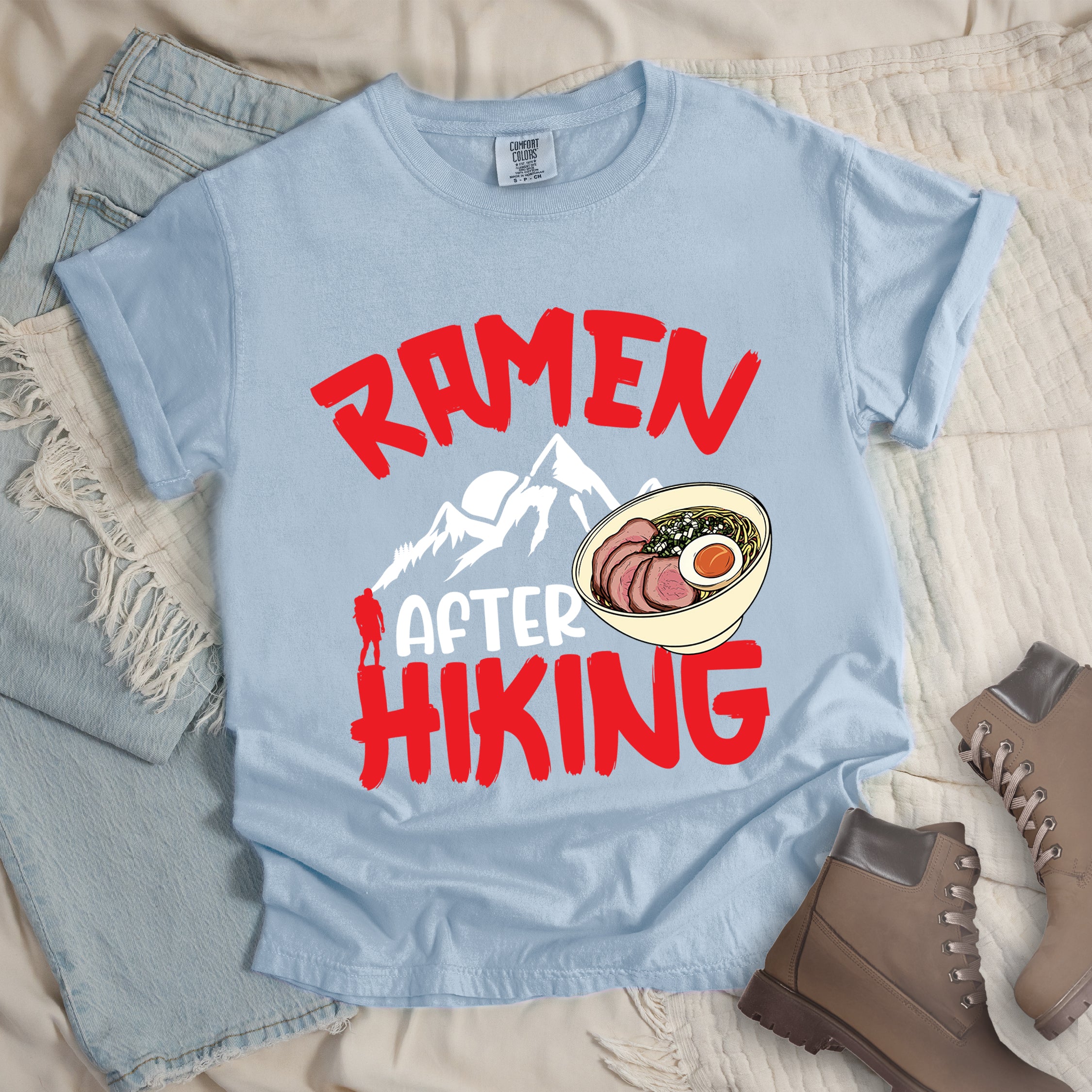 Chambray (Light Blue) t-shirt with red text reading "RAMEN AFTER HIKING". "RAMEN" appears in bold red letters at the top, "AFTER" in white in the middle, and "HIKING" in bold red at the bottom. Features white mountain silhouettes in the center with a red hiker figure, and a detailed illustration of a ramen bowl with sliced meat, egg, and green onions on the right. The design connects outdoor activities with food rewards.