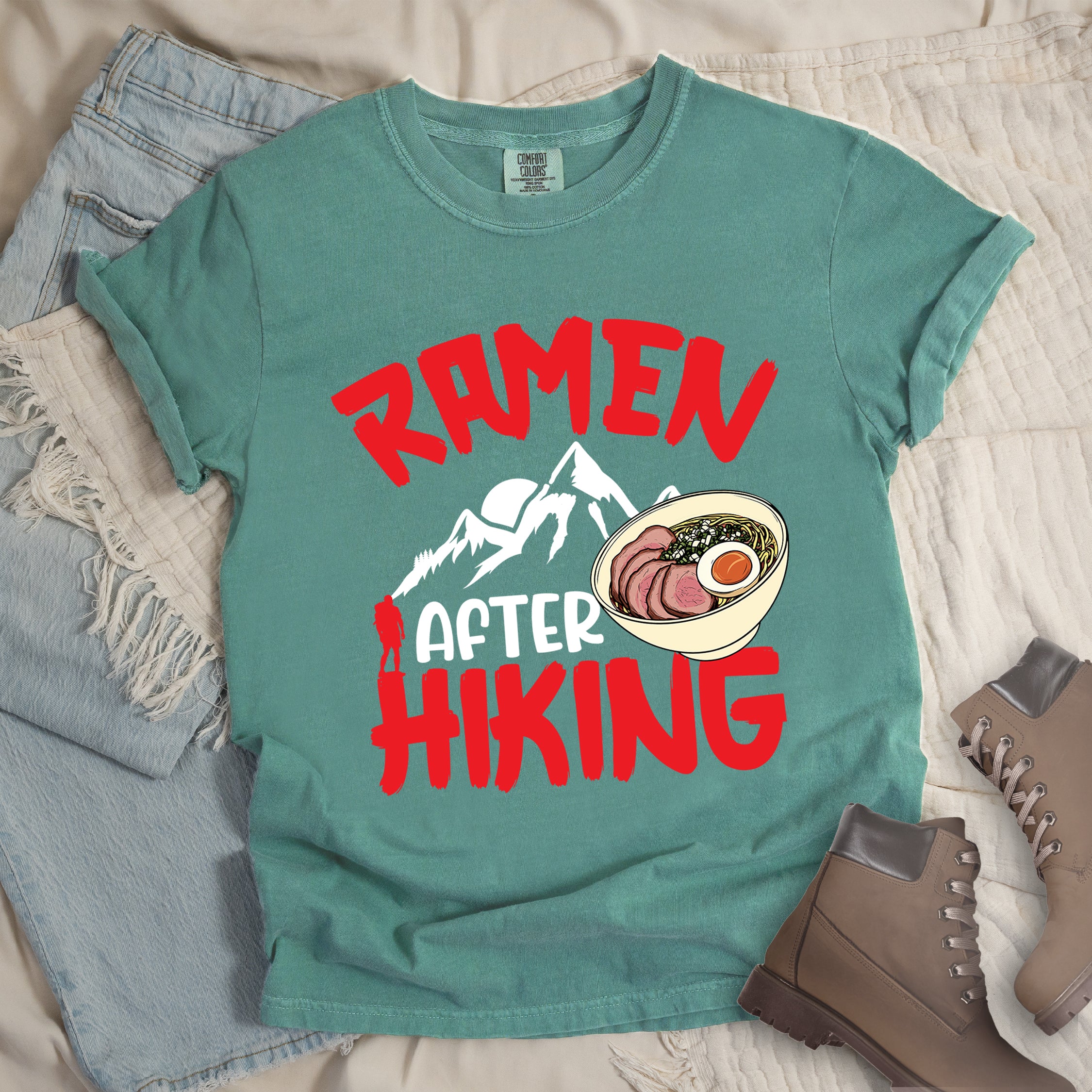 Light Green t-shirt with red text reading "RAMEN AFTER HIKING". "RAMEN" appears in bold red letters at the top, "AFTER" in white in the middle, and "HIKING" in bold red at the bottom. Features white mountain silhouettes in the center with a red hiker figure, and a detailed illustration of a ramen bowl with sliced meat, egg, and green onions on the right. The design connects outdoor activities with food rewards.