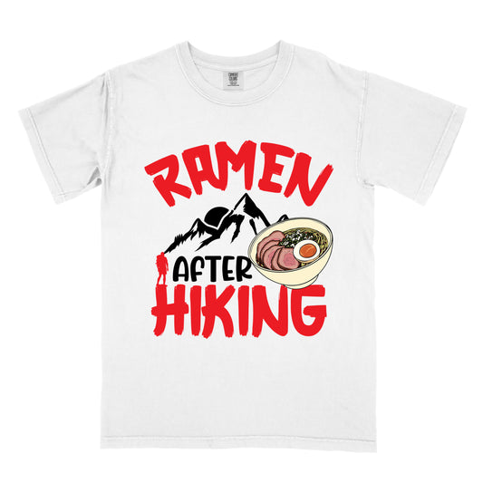 White t-shirt with red text reading "RAMEN AFTER HIKING". "RAMEN" appears in bold red letters at the top, "AFTER" in black in the middle, and "HIKING" in bold red at the bottom. Features black mountain silhouettes in the center with a red hiker figure, and a detailed illustration of a ramen bowl with sliced meat, egg, and green onions on the right. The design connects outdoor activities with food rewards.