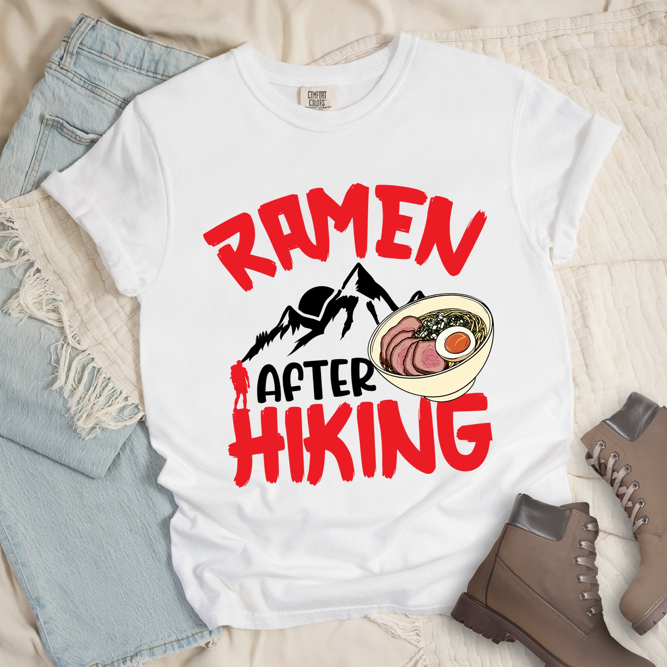 White t-shirt with red text reading "RAMEN AFTER HIKING". "RAMEN" appears in bold red letters at the top, "AFTER" in black in the middle, and "HIKING" in bold red at the bottom. Features black mountain silhouettes in the center with a red hiker figure, and a detailed illustration of a ramen bowl with sliced meat, egg, and green onions on the right. The design connects outdoor activities with food rewards. 