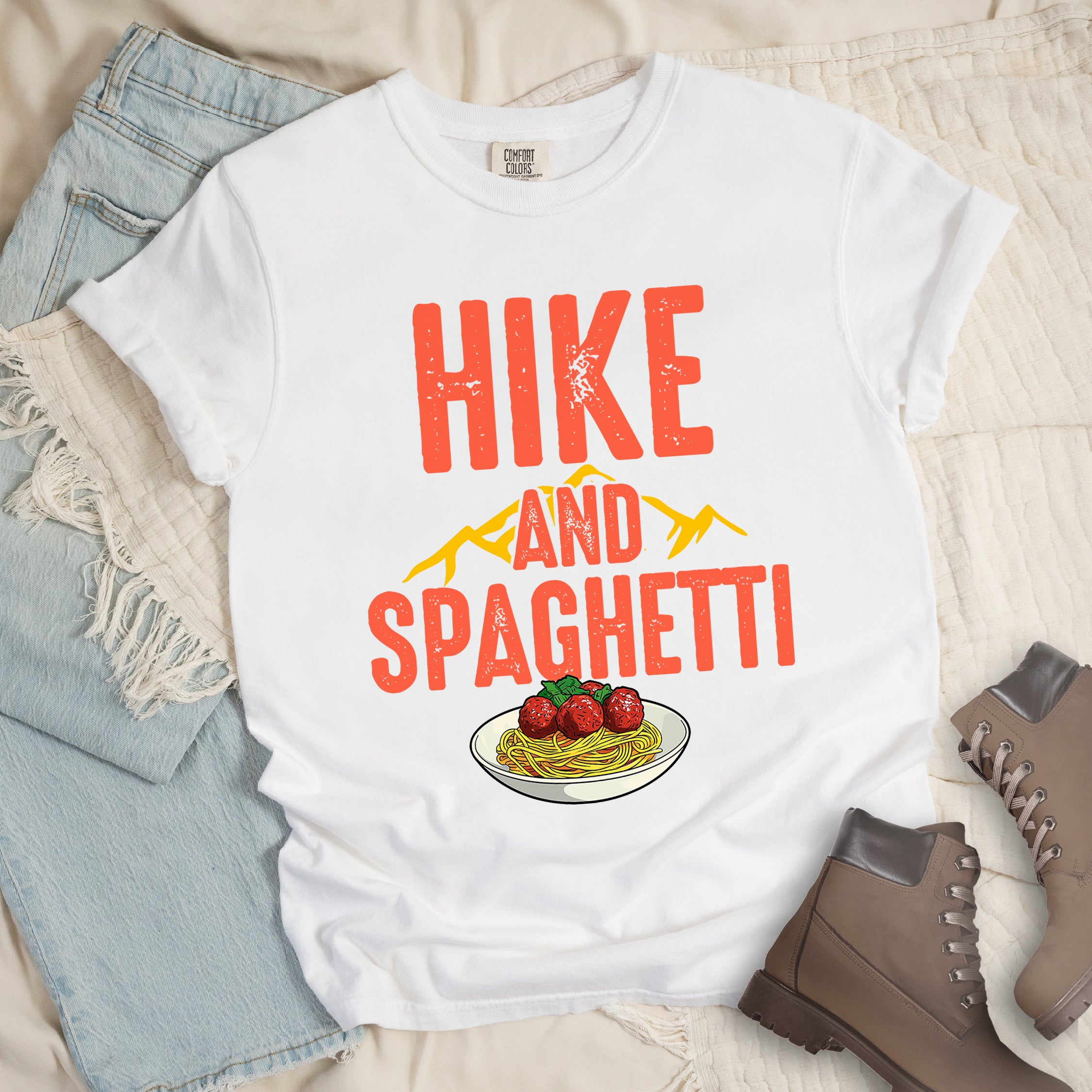 White t-shirt with red distressed text reading "HIKE AND SPAGHETTI". "HIKE" appears in large red letters at the top, "AND" in red over yellow mountain silhouettes in the middle, and "SPAGHETTI" in large red letters at the bottom. Features a detailed illustration of a white plate with spaghetti, meatballs, and herbs at the bottom of the design.Features a classic crew neck and casual rolled sleeves, styled alongside light wash denim and brown hiking boots. 