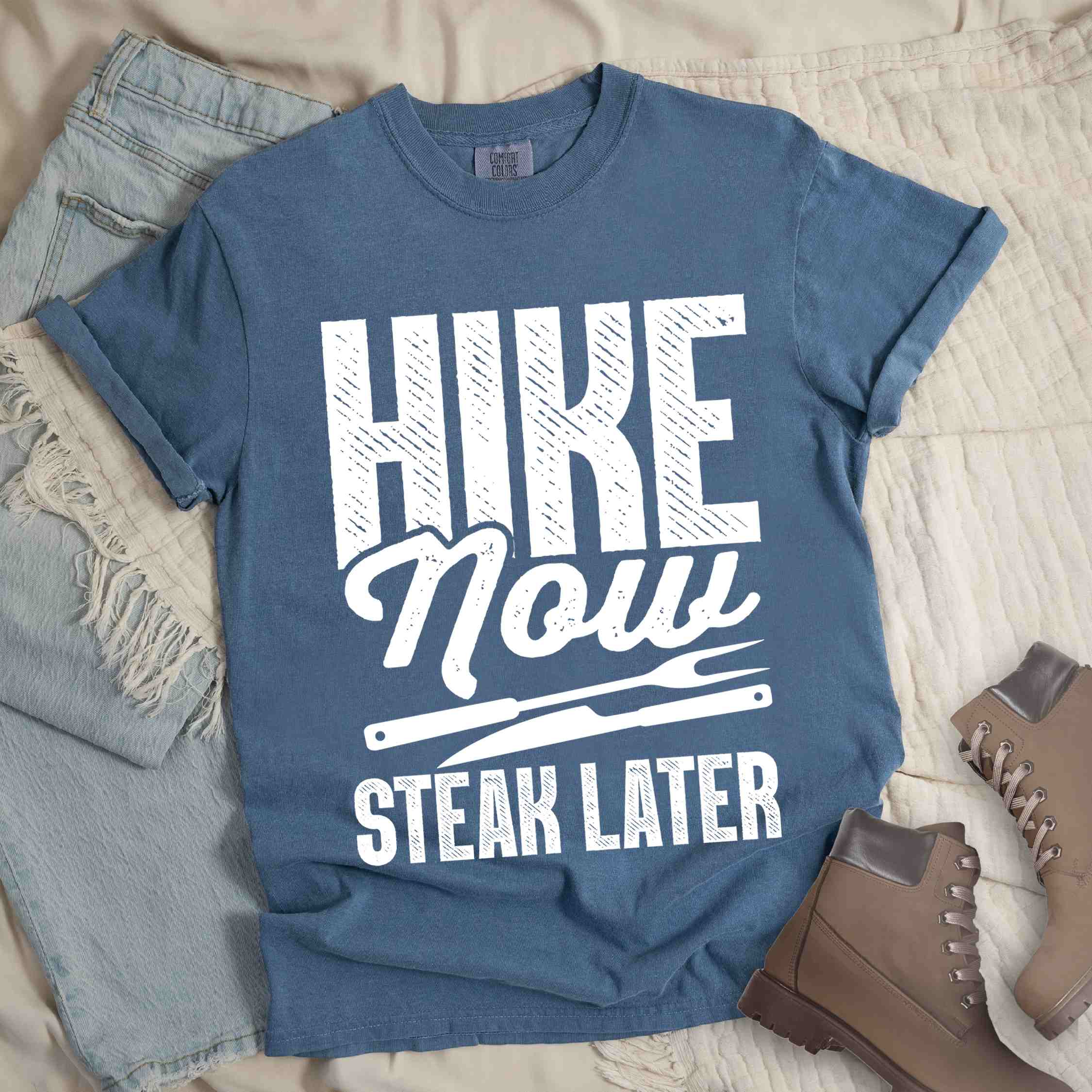 Blue jean "Hike now steak later" colored vintage tee with bold typography, illustrated steak utensils, and backpacker for steak lovers.