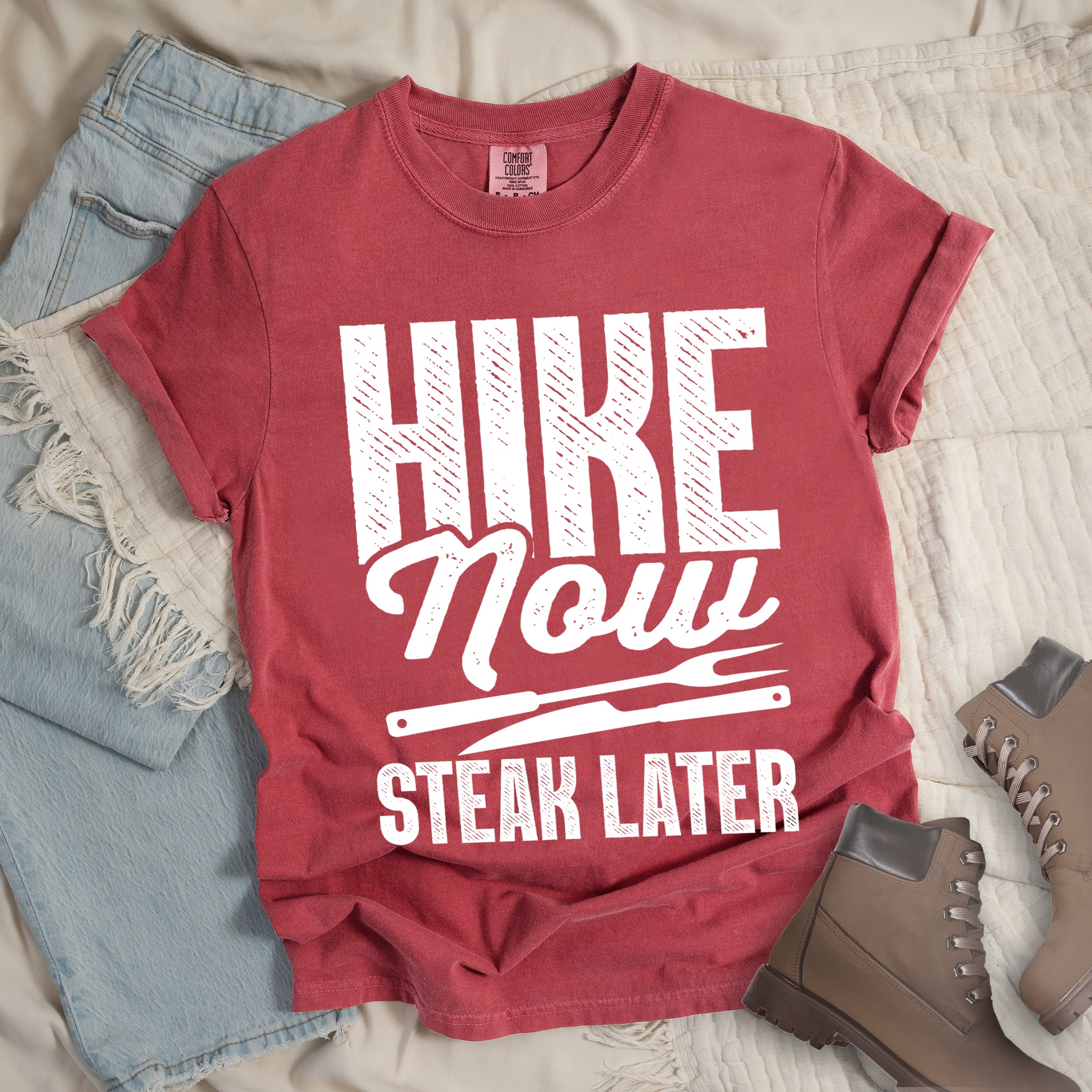 Crimson (Dark Pink) t-shirt with white distressed text reading "HIKE Now STEAK LATER". "HIKE" appears in large bold block letters at the top, "Now" in stylish script in the middle, and "STEAK LATER" in block letters at the bottom. Between the middle and bottom text is a simple illustration of a steak knife. The design emphasizes the reward of enjoying steak after completing a hiking activity. Features a classic crew neck and casual rolled sleeves, styled alongside light wash denim and brown hiking boots. 