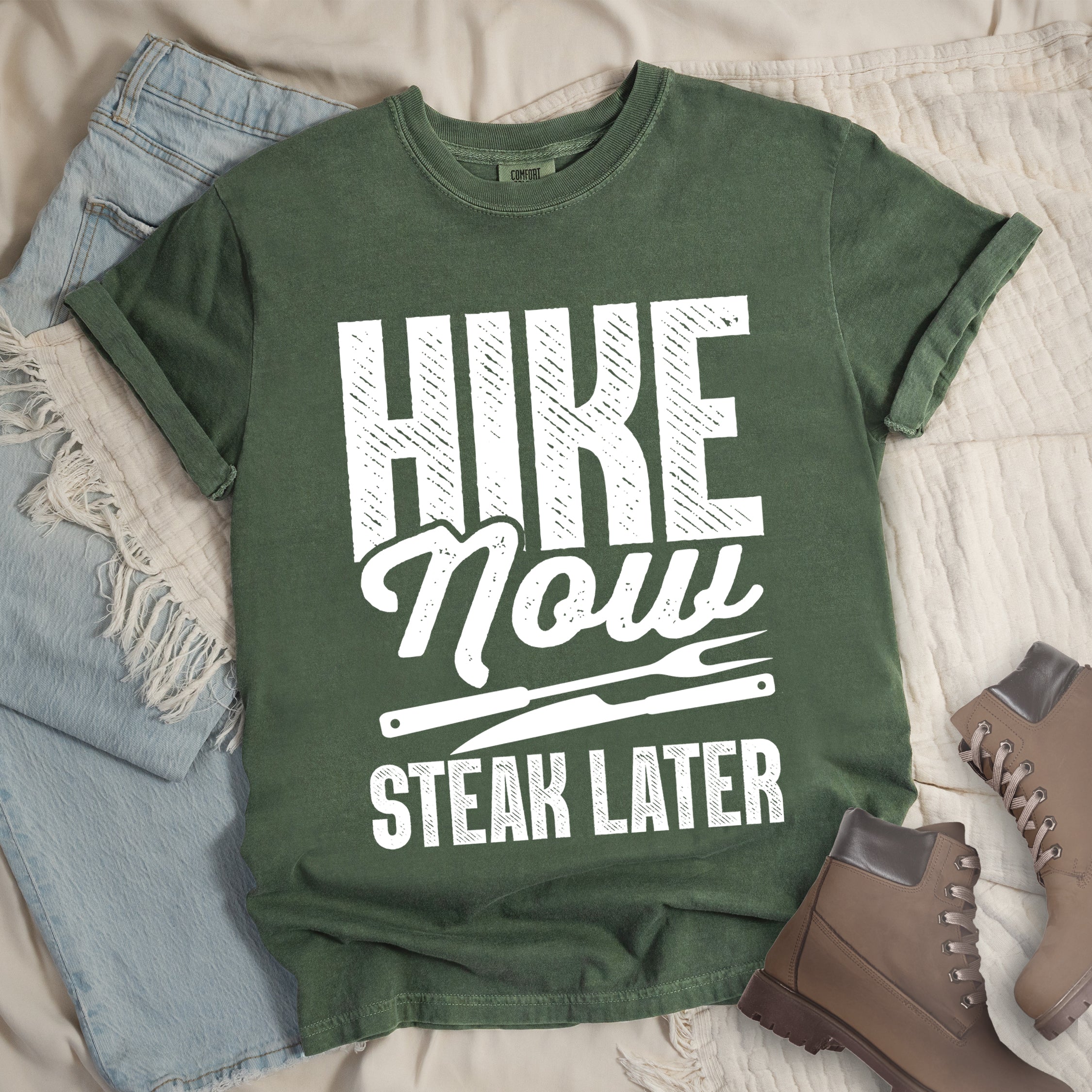 Moss (Dark Green) t-shirt with white distressed text reading "HIKE Now STEAK LATER". "HIKE" appears in large bold block letters at the top, "Now" in stylish script in the middle, and "STEAK LATER" in block letters at the bottom. Between the middle and bottom text is a simple illustration of a steak knife. The design emphasizes the reward of enjoying steak after completing a hiking activity. Features a classic crew neck and casual rolled sleeves, styled alongside light wash denim and brown hiking boots. 