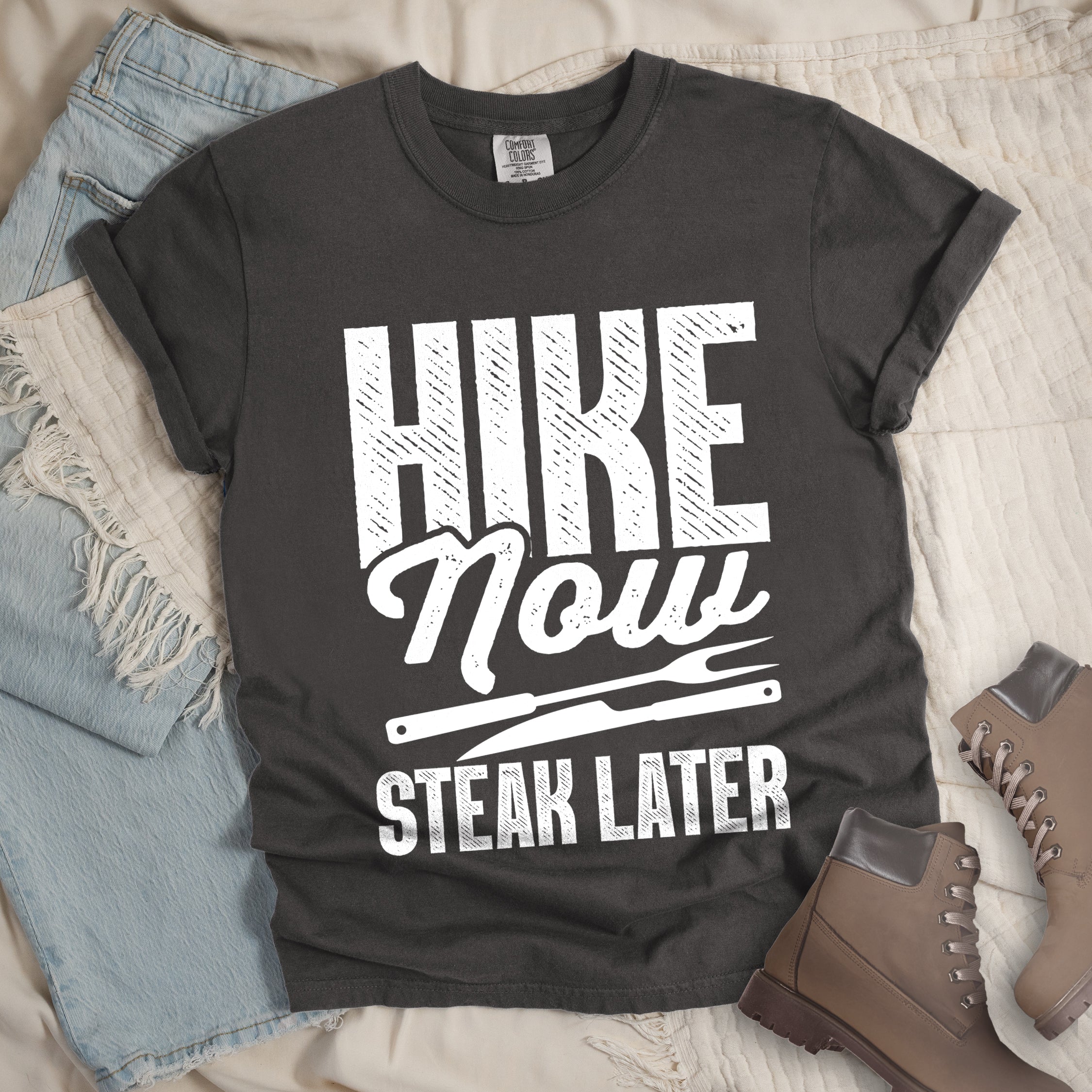 Pepper (Dark Gray) t-shirt with white distressed text reading "HIKE Now STEAK LATER". "HIKE" appears in large bold block letters at the top, "Now" in stylish script in the middle, and "STEAK LATER" in block letters at the bottom. Between the middle and bottom text is a simple illustration of a steak knife. The design emphasizes the reward of enjoying steak after completing a hiking activity. Features a classic crew neck and casual rolled sleeves, styled alongside light wash denim and brown hiking boots. 