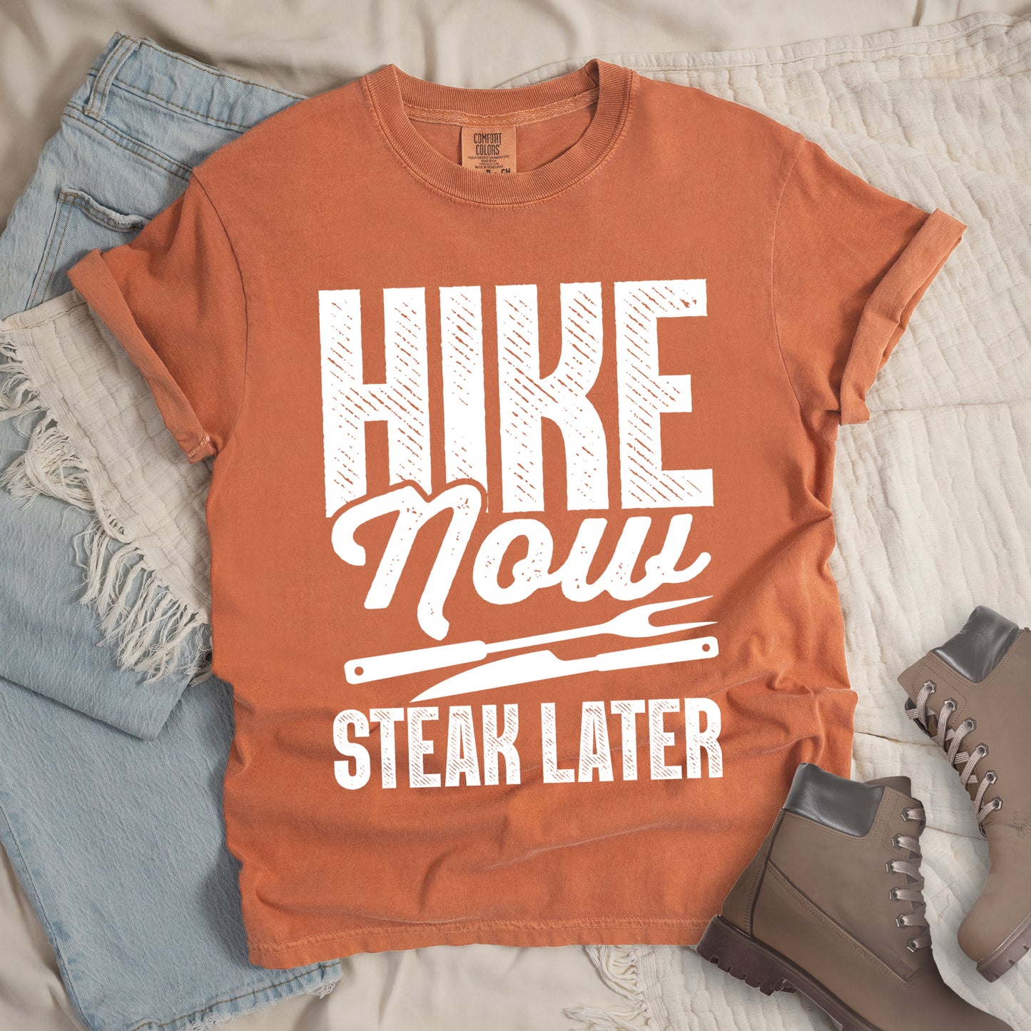 Yam (Brown) t-shirt with white distressed text reading "HIKE Now STEAK LATER". "HIKE" appears in large bold block letters at the top, "Now" in stylish script in the middle, and "STEAK LATER" in block letters at the bottom. Between the middle and bottom text is a simple illustration of a steak knife. The design emphasizes the reward of enjoying steak after completing a hiking activity. Features a classic crew neck and casual rolled sleeves, styled alongside light wash denim and brown hiking boots. 