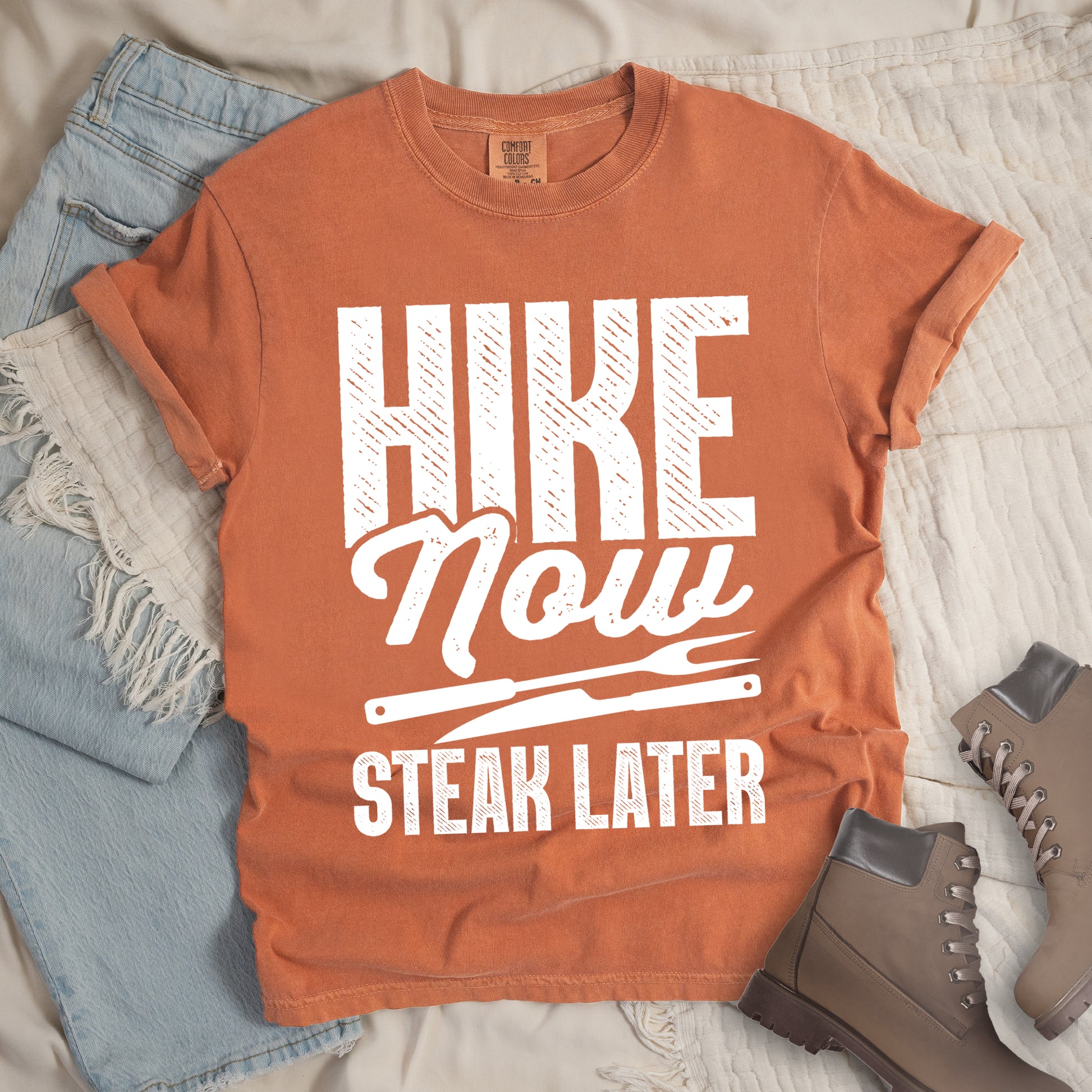 Yam (Brown) t-shirt with white distressed text reading "HIKE Now STEAK LATER". "HIKE" appears in large bold block letters at the top, "Now" in stylish script in the middle, and "STEAK LATER" in block letters at the bottom. Between the middle and bottom text is a simple illustration of a steak knife. The design emphasizes the reward of enjoying steak after completing a hiking activity. Features a classic crew neck and casual rolled sleeves, styled alongside light wash denim and brown hiking boots. 