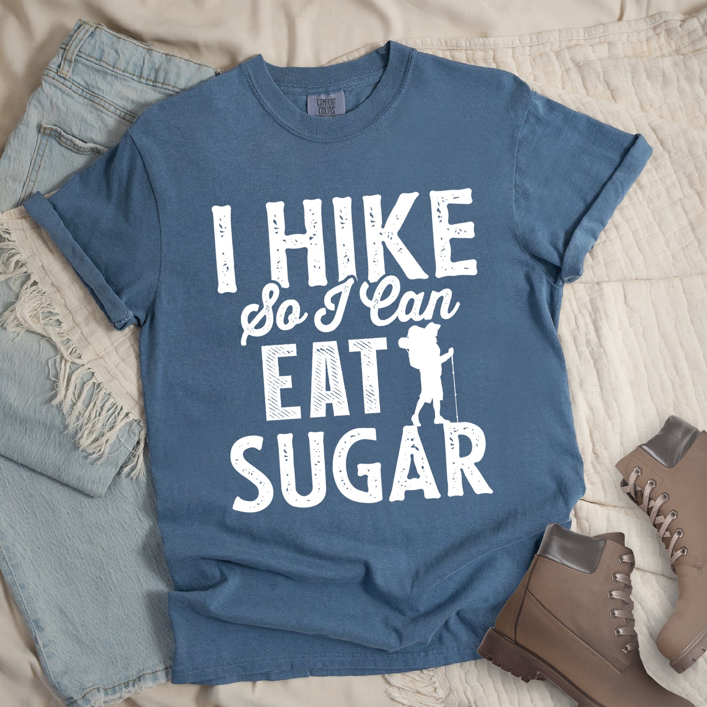 Blue Jean (Denim Blue) t-shirt with white distressed text reading "I HIKE So I Can EAT SUGAR". "I HIKE" appears in large block letters at the top, "So I Can" in script in the middle, and "EAT SUGAR" in block letters at the bottom. A small silhouette of a hiker with backpack and walking stick is positioned next to the word "EAT" and above "SUGAR". Features a classic crew neck and casual rolled sleeves, styled alongside light wash denim and brown hiking boots. 