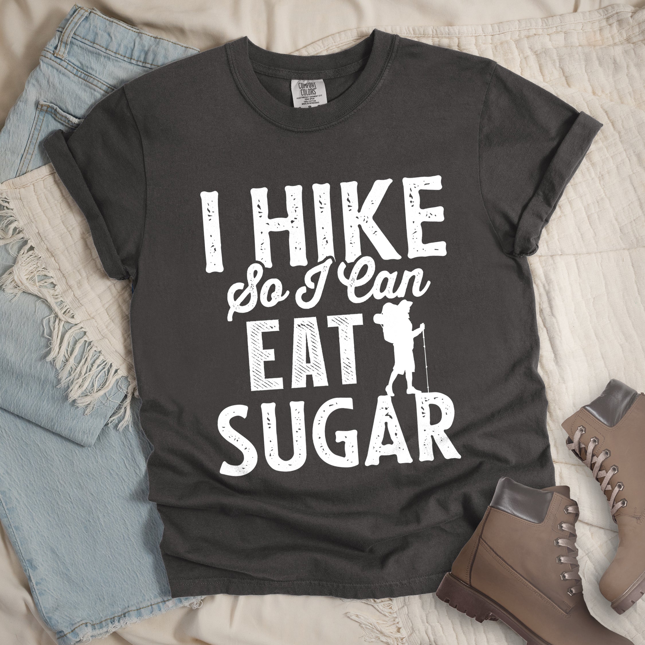 Pepper (Dark Gray) t-shirt with white distressed text reading "I HIKE So I Can EAT SUGAR". "I HIKE" appears in large block letters at the top, "So I Can" in script in the middle, and "EAT SUGAR" in block letters at the bottom. A small silhouette of a hiker with backpack and walking stick is positioned next to the word "EAT" and above "SUGAR". Features a classic crew neck and casual rolled sleeves, styled alongside light wash denim and brown hiking boots. 