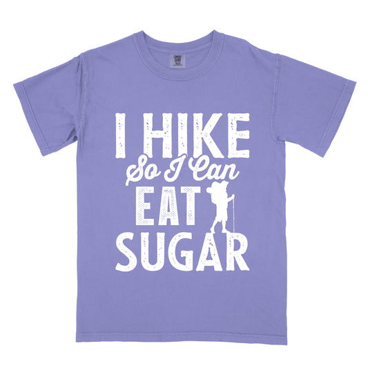 Violet t-shirt with white distressed text reading "I HIKE So I Can EAT SUGAR". "I HIKE" appears in large block letters at the top, "So I Can" in script in the middle, and "EAT SUGAR" in block letters at the bottom. A small silhouette of a hiker with backpack and walking stick is positioned next to the word "EAT" and above "SUGAR".