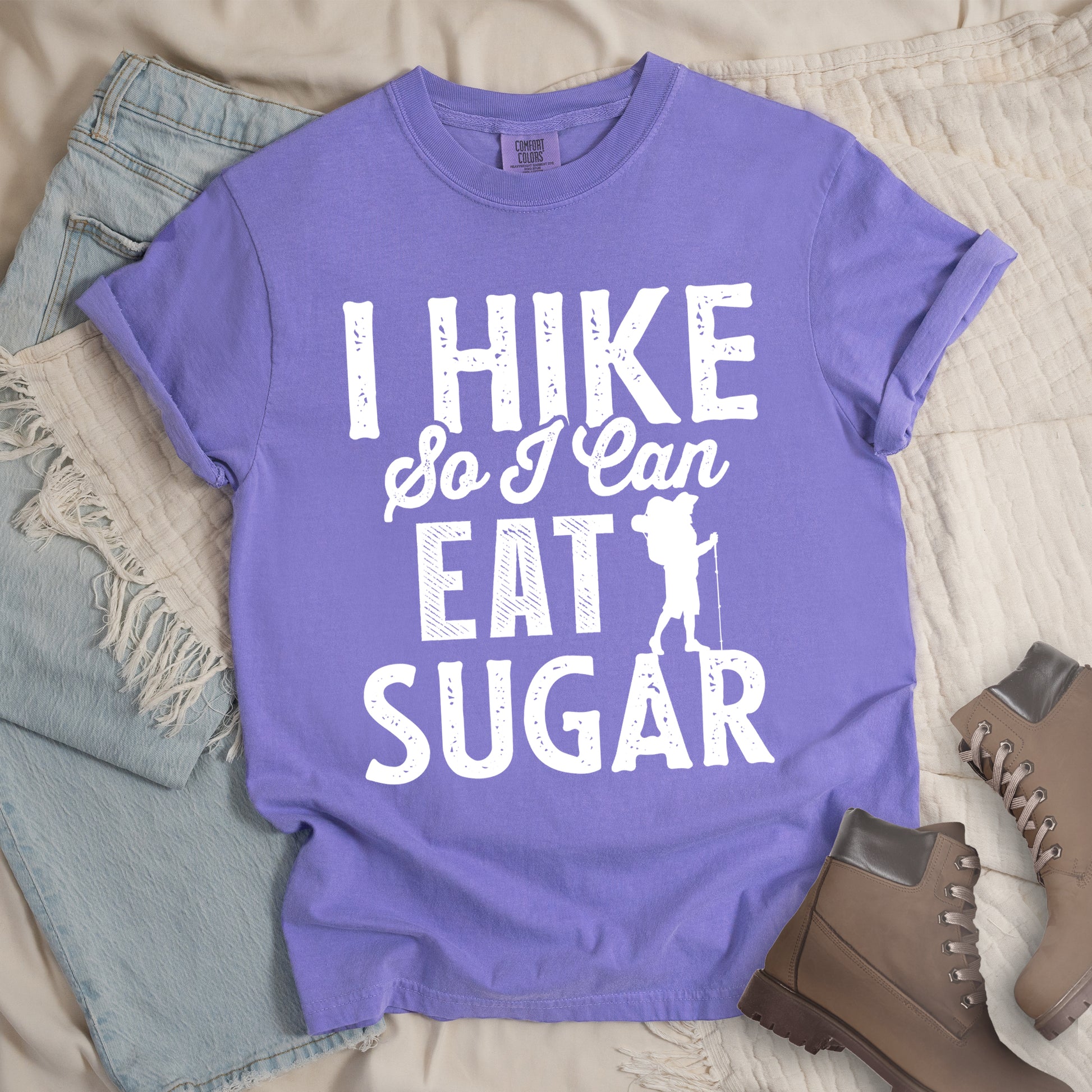 Violet t-shirt with white distressed text reading "I HIKE So I Can EAT SUGAR". "I HIKE" appears in large block letters at the top, "So I Can" in script in the middle, and "EAT SUGAR" in block letters at the bottom. A small silhouette of a hiker with backpack and walking stick is positioned next to the word "EAT" and above "SUGAR". Features a classic crew neck and casual rolled sleeves, styled alongside light wash denim and brown hiking boots. 