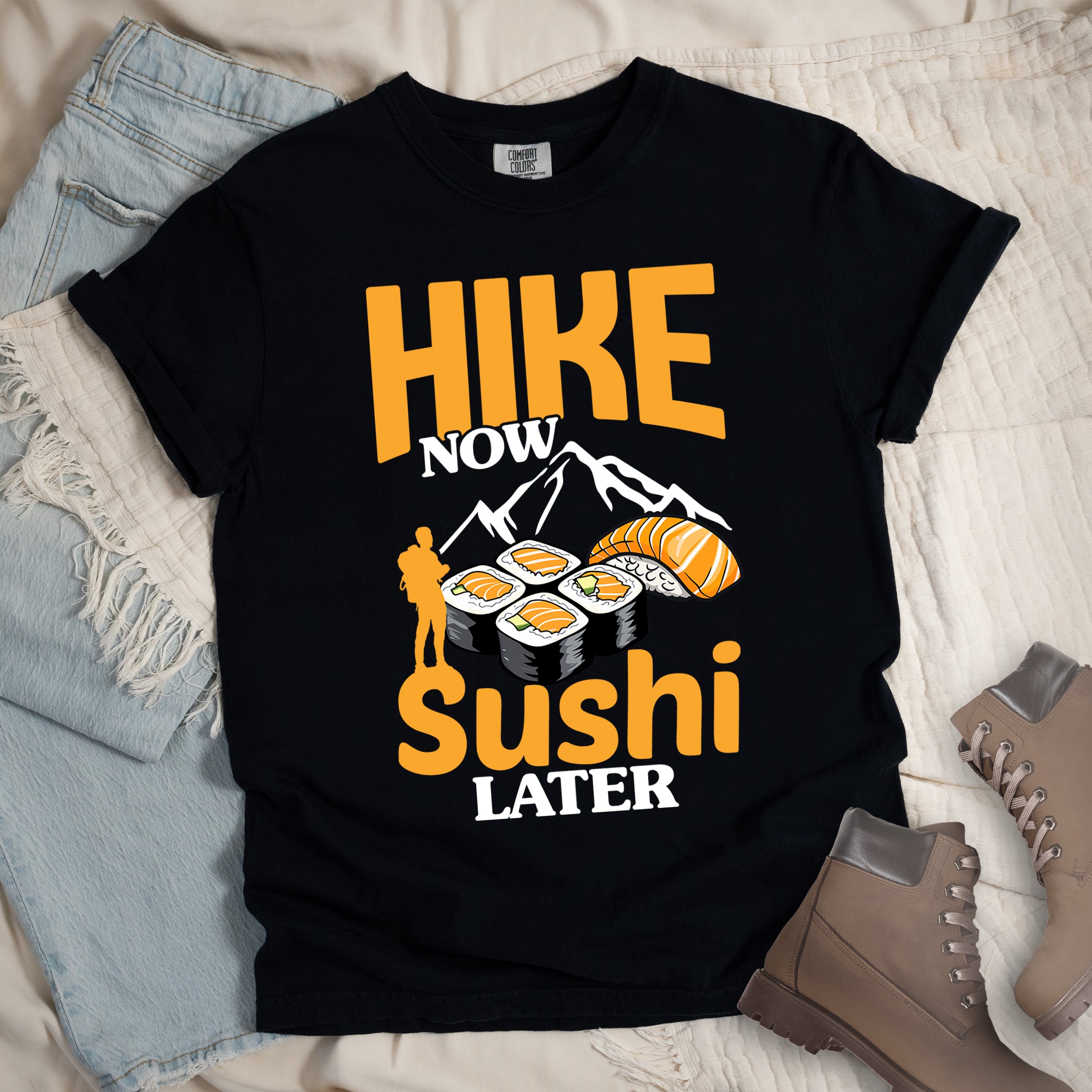 Black t-shirt with orange and white text reading "HIKE NOW sushi LATER". "HIKE" appears in large orange letters at the top, "NOW" in white, "sushi" in orange at the bottom, and "LATER" in white. Features white mountain silhouettes in the center, detailed sushi illustrations including rolls and nigiri in orange/white, and an orange silhouette of a hiker with backpack on the left. 