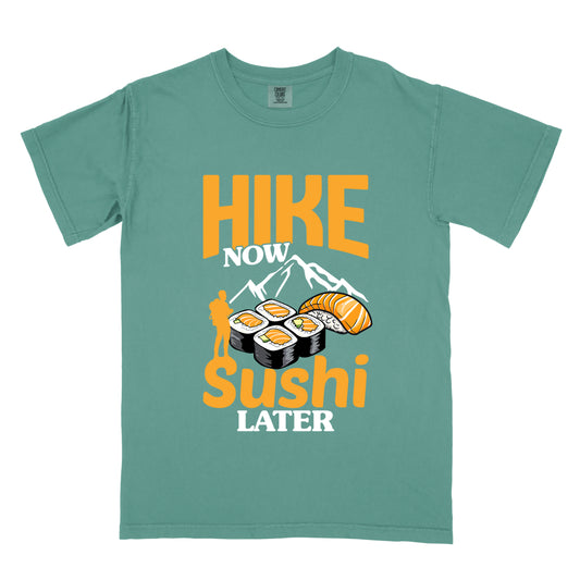 Light Green t-shirt with orange and white text reading "HIKE NOW sushi LATER". "HIKE" appears in large orange letters at the top, "NOW" in white, "sushi" in orange at the bottom, and "LATER" in white. Features white mountain silhouettes in the center, detailed sushi illustrations including rolls and nigiri in orange/white, and an orange silhouette of a hiker with backpack on the left. 