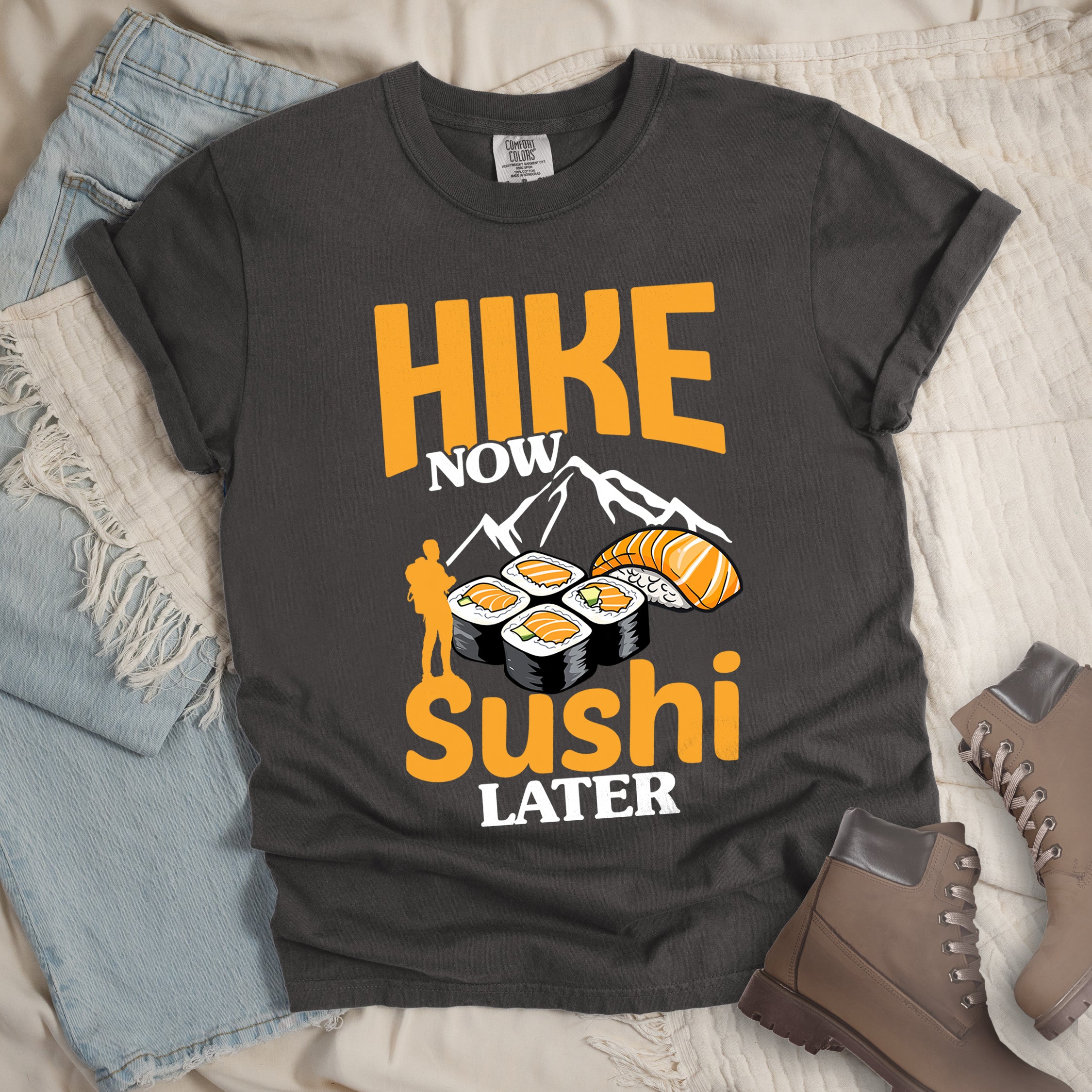 Pepper (Dark Gray) t-shirt with orange and white text reading "HIKE NOW sushi LATER". "HIKE" appears in large orange letters at the top, "NOW" in white, "sushi" in orange at the bottom, and "LATER" in white. Features white mountain silhouettes in the center, detailed sushi illustrations including rolls and nigiri in orange/white, and an orange silhouette of a hiker with backpack on the left. 