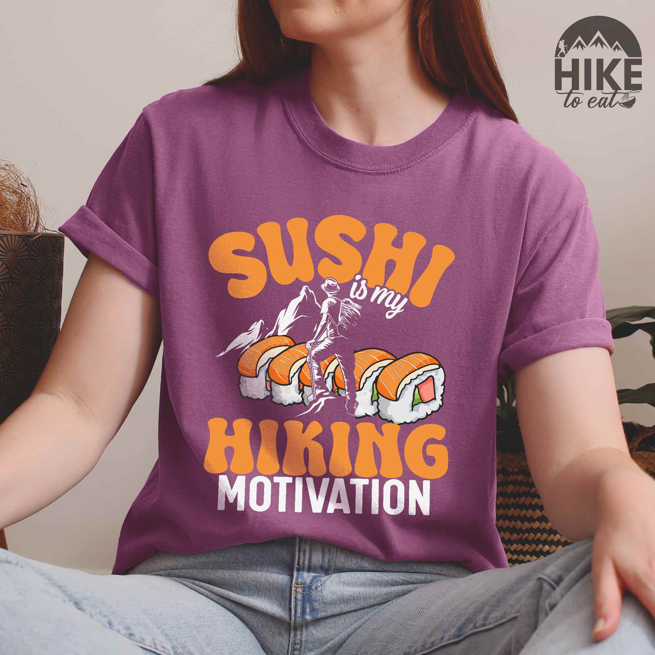Woman wearing berry "sushi is my hiking motivation" colored vintage tee with bold typography, birds, mountains, illustrated sushi platter, and backpacker for sushi lovers.