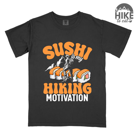 Black t-shirt with orange and white text reading "SUSHI is my HIKING MOTIVATION". "SUSHI" appears in large orange letters at the top, "is my" in small white text, and "HIKING" in orange with "MOTIVATION" in white at the bottom. Features a humorous illustration of a hiker climbing over giant sushi rolls, with white mountain silhouettes in the background. 