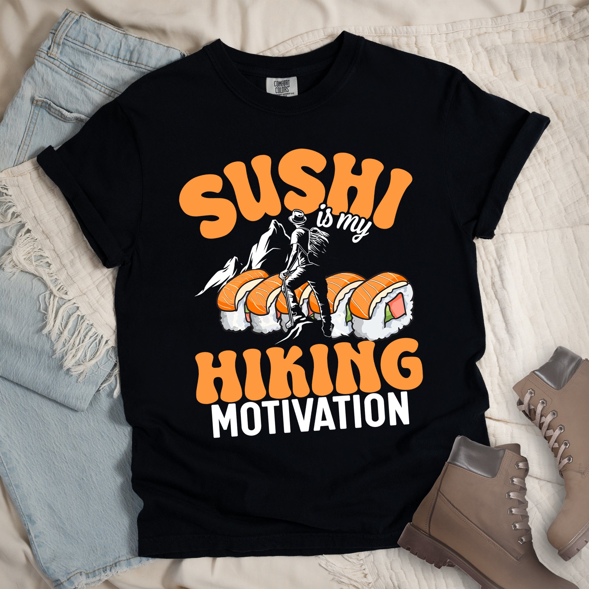 Black t-shirt with orange and white text reading "SUSHI is my HIKING MOTIVATION". "SUSHI" appears in large orange letters at the top, "is my" in small white text, and "HIKING" in orange with "MOTIVATION" in white at the bottom. Features a humorous illustration of a hiker climbing over giant sushi rolls, with white mountain silhouettes in the background. 