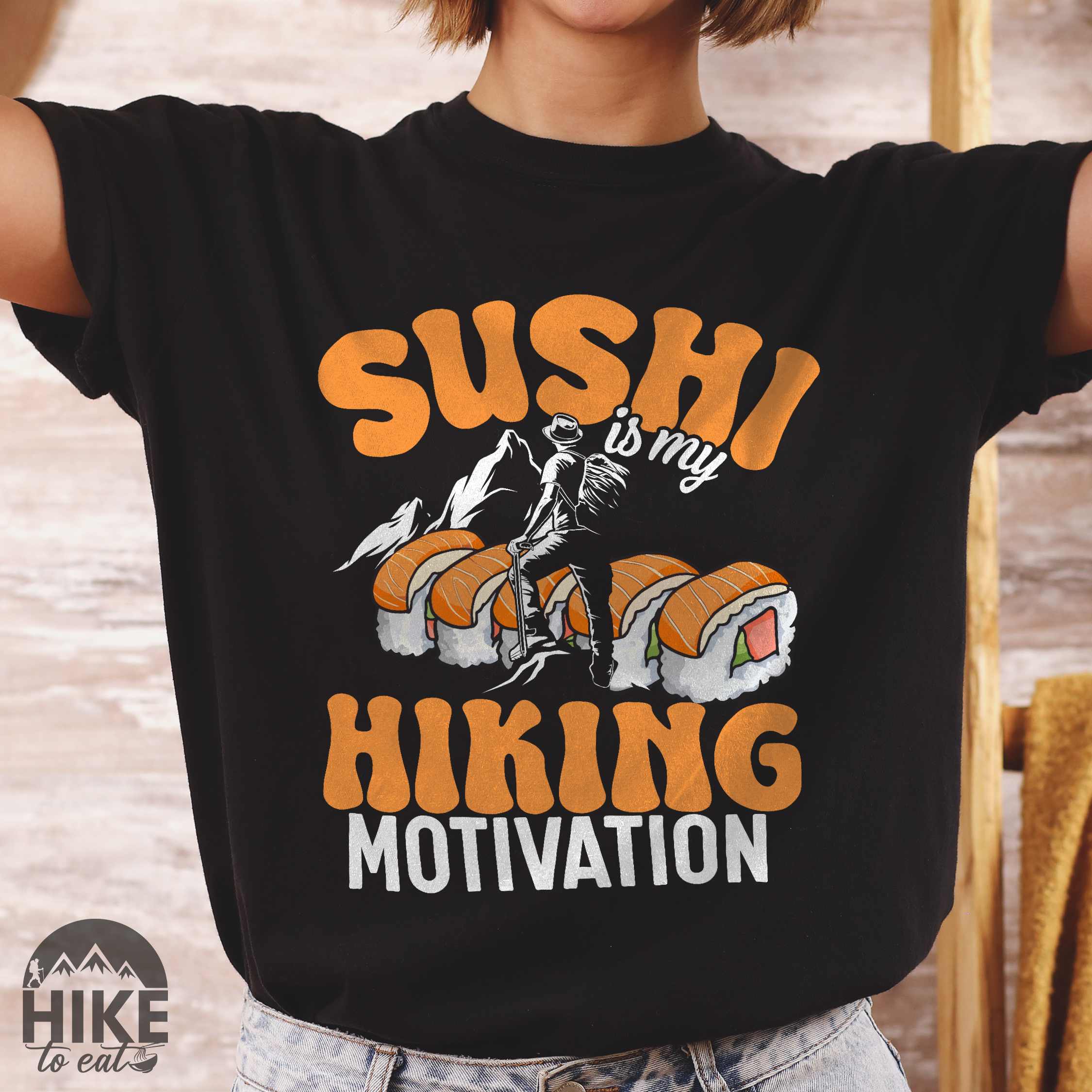 Woman wearing black "sushi is my hiking motivation" colored vintage tee with bold typography, birds, mountains, illustrated sushi platter, and backpacker for sushi lovers.