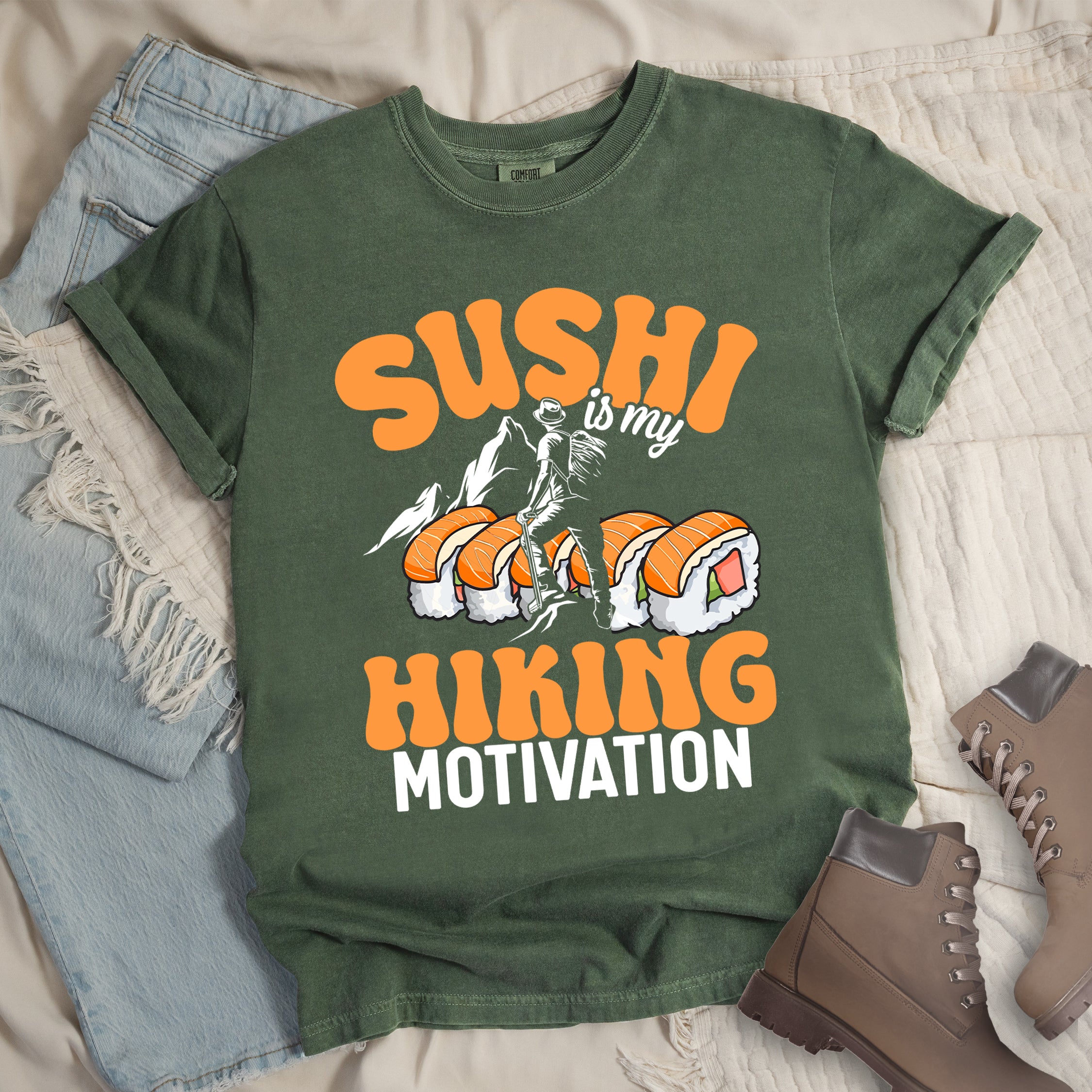 Moss (Dark Green) t-shirt with orange and white text reading "SUSHI is my HIKING MOTIVATION". "SUSHI" appears in large orange letters at the top, "is my" in small white text, and "HIKING" in orange with "MOTIVATION" in white at the bottom. Features a humorous illustration of a hiker climbing over giant sushi rolls, with white mountain silhouettes in the background. 