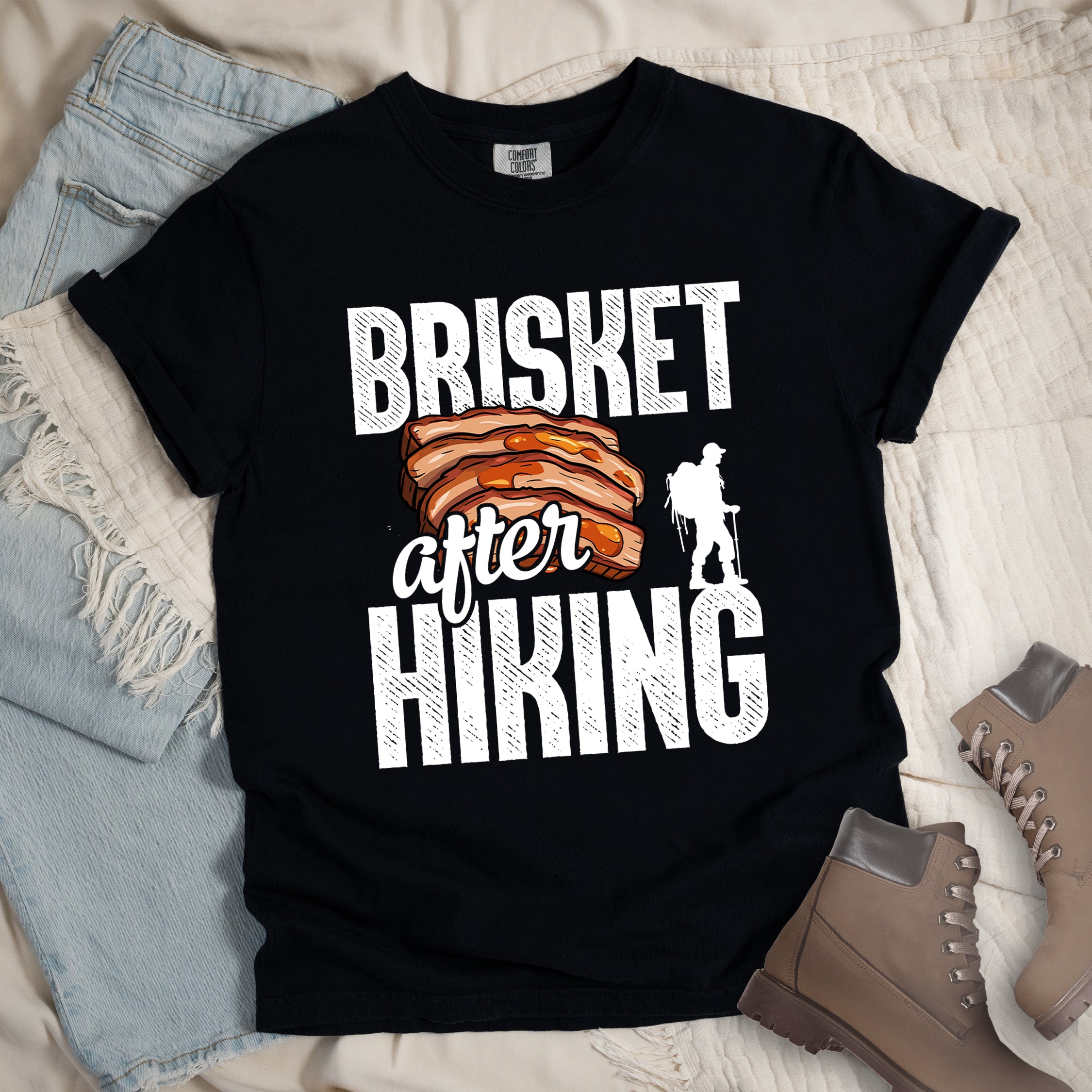 Black t-shirt with white distressed text reading "BRISKET after HIKING". "BRISKET" appears in large white letters at the top, "after" in white script in the middle, and "HIKING" in large white letters at the bottom. Features a detailed illustration of sliced brisket meat in orange-brown tones in the center and a white silhouette of a hiker with walking poles on the right side.