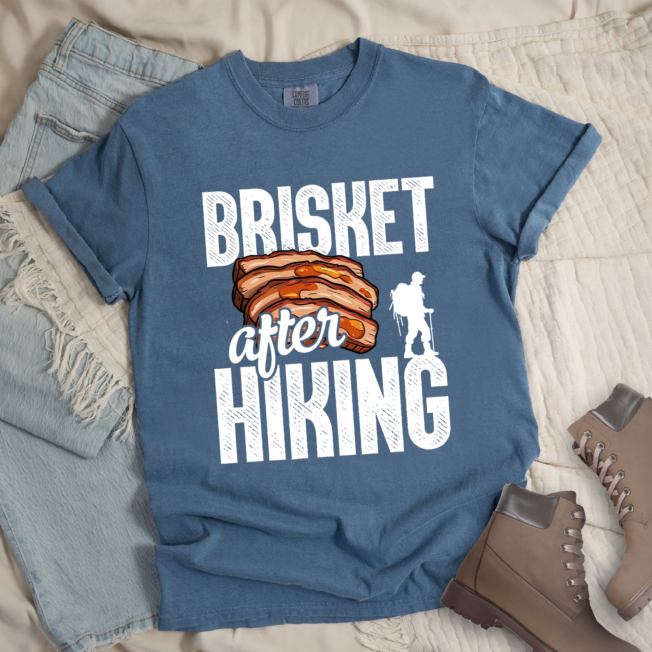 Blue Jean (Denim Blue) t-shirt with white distressed text reading "BRISKET after HIKING". "BRISKET" appears in large white letters at the top, "after" in white script in the middle, and "HIKING" in large white letters at the bottom. Features a detailed illustration of sliced brisket meat in orange-brown tones in the center and a white silhouette of a hiker with walking poles on the right side.