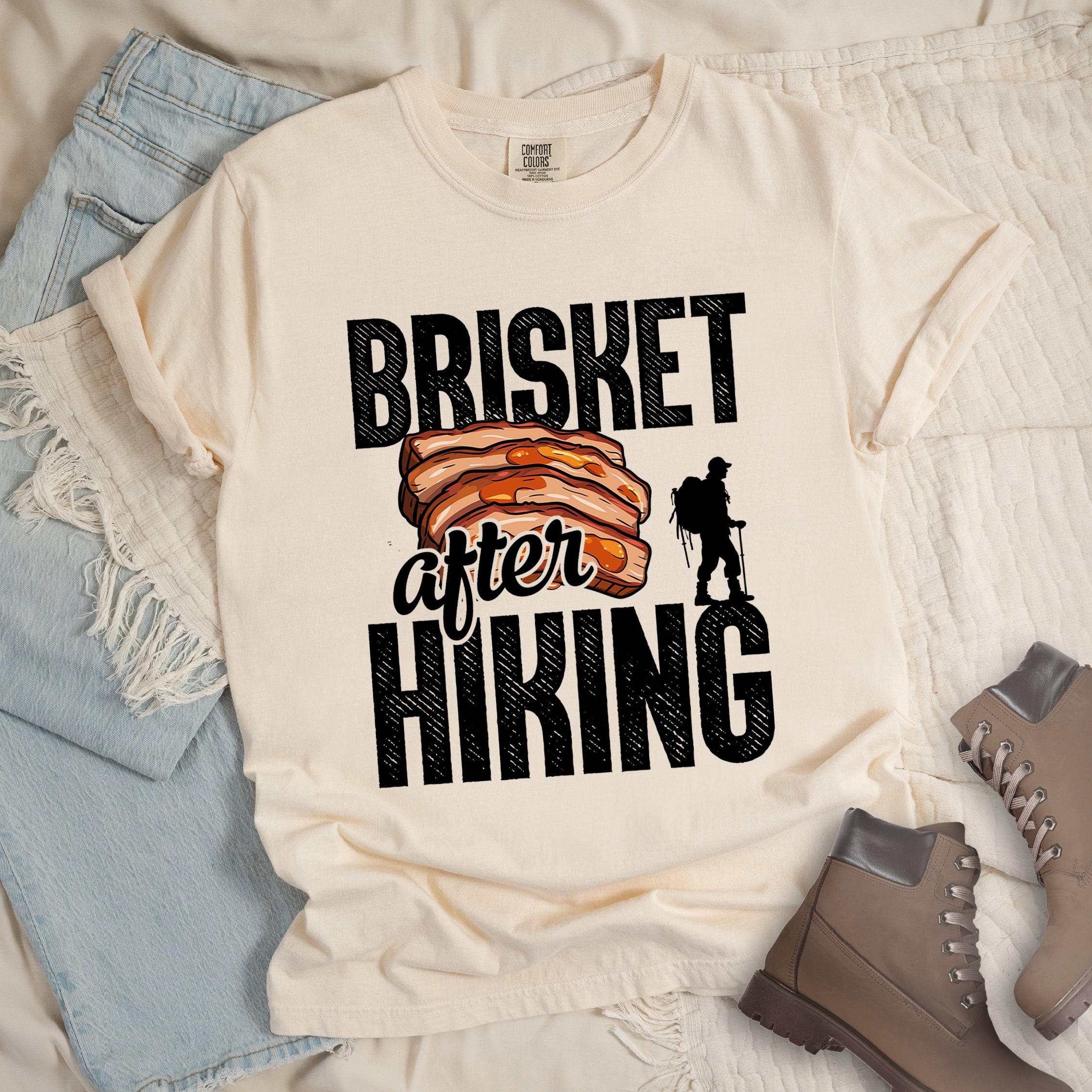 Ivory (Cream White) t-shirt with black distressed text reading "BRISKET after HIKING". "BRISKET" appears in large black letters at the top, "after" in black script in the middle, and "HIKING" in large black letters at the bottom. Features a detailed illustration of sliced brisket meat in orange-brown tones in the center and a black silhouette of a hiker with walking poles on the right side.