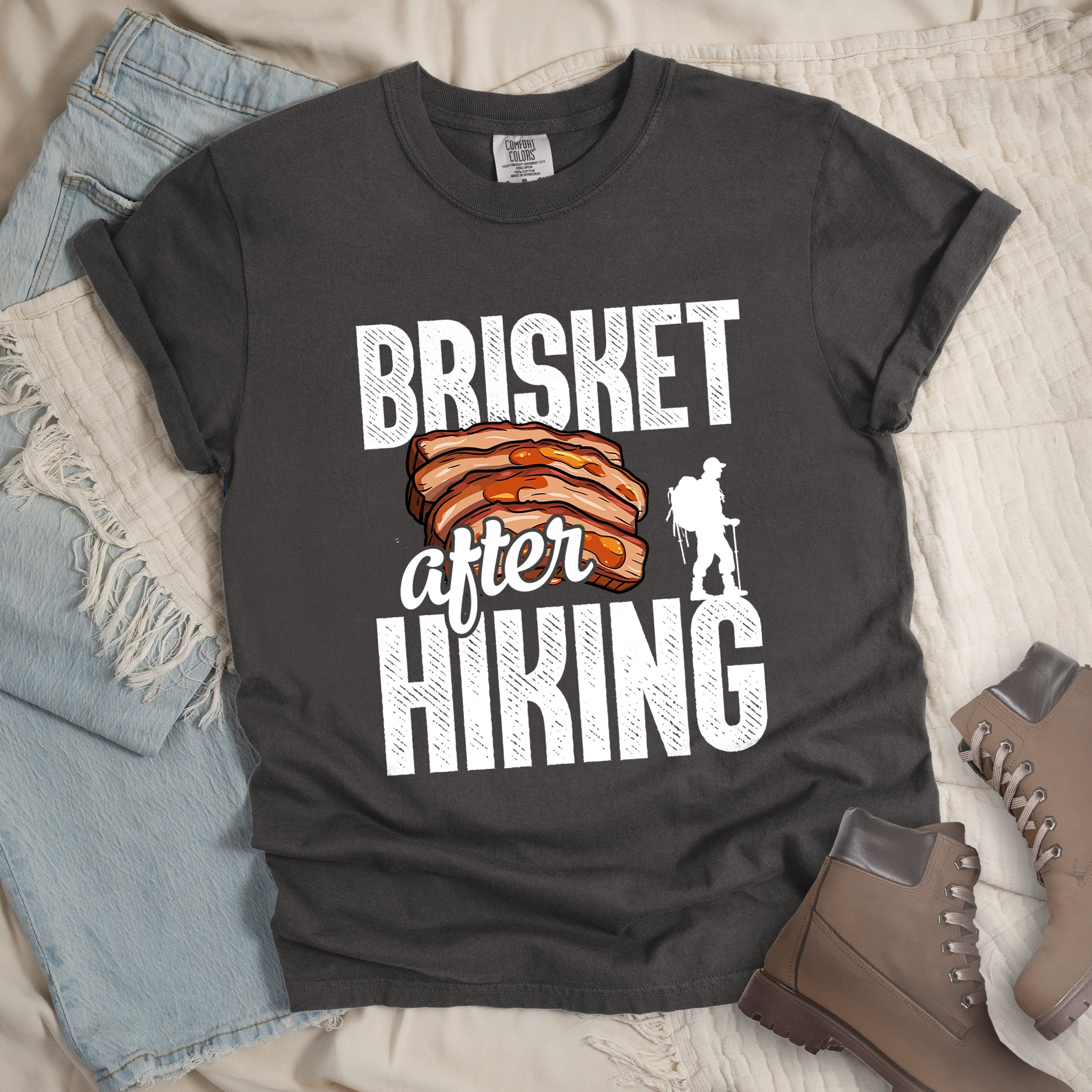 Pepper (Dark Gray) t-shirt with white distressed text reading "BRISKET after HIKING". "BRISKET" appears in large white letters at the top, "after" in white script in the middle, and "HIKING" in large white letters at the bottom. Features a detailed illustration of sliced brisket meat in orange-brown tones in the center and a white silhouette of a hiker with walking poles on the right side.